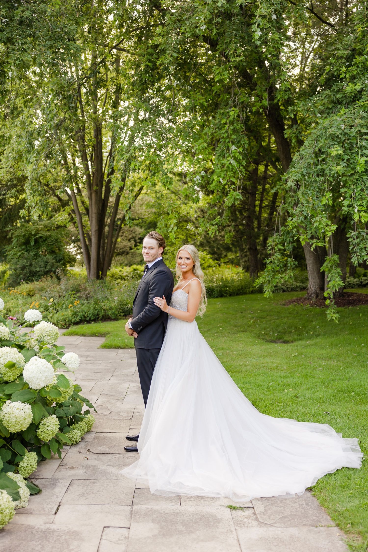Couple poses back to back on garden path surrounded by lush trees and white hydrangeas.