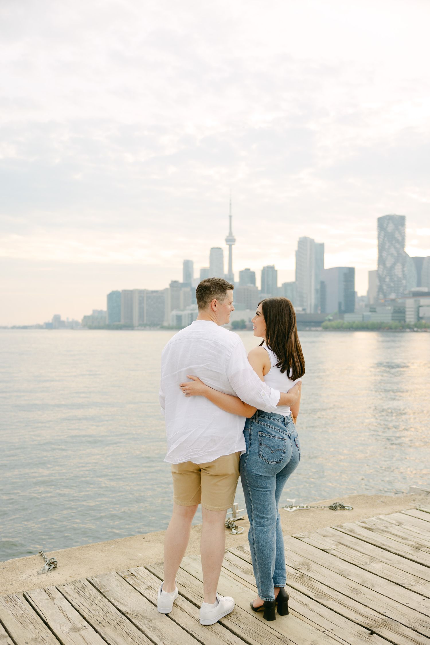 A romantic couple embraces on a wooden pier with the Toronto skyline visible across the water.