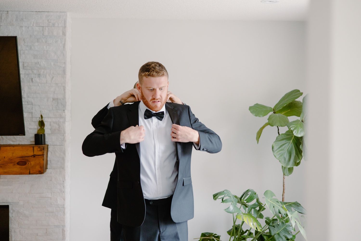 Person in formal attire adjusts their suit jacket in a modern room with houseplants.