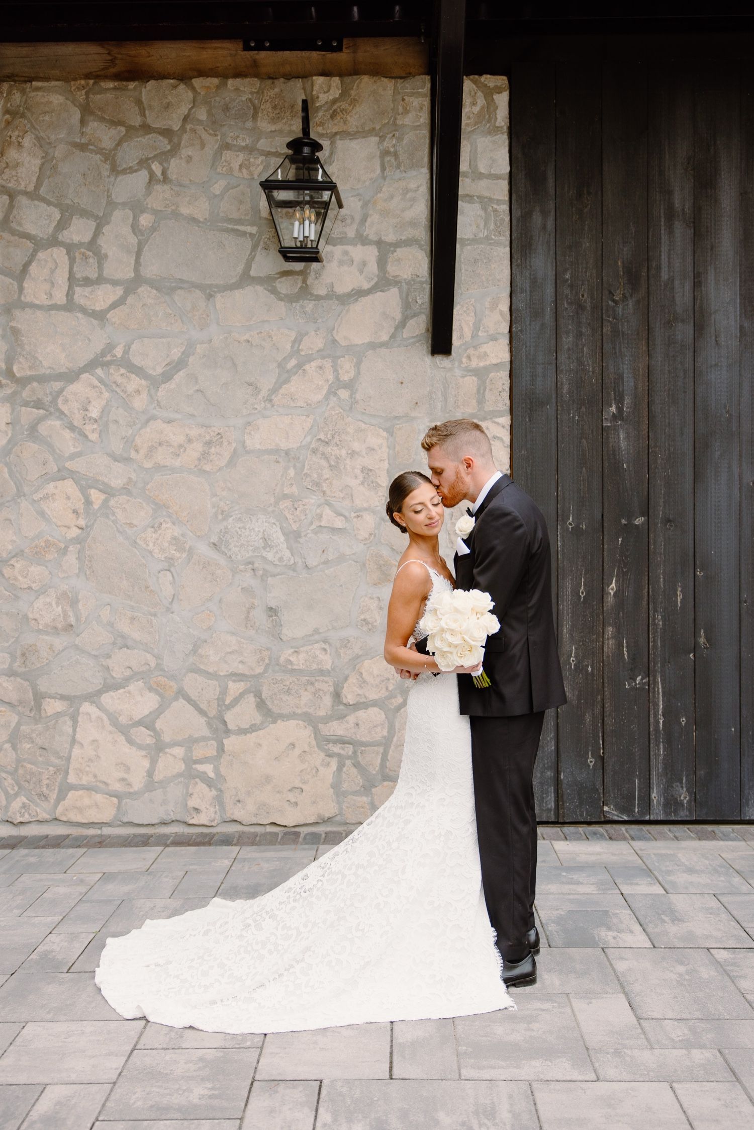 A couple shares an intimate moment against a stone wall with a rustic lantern and wooden door.