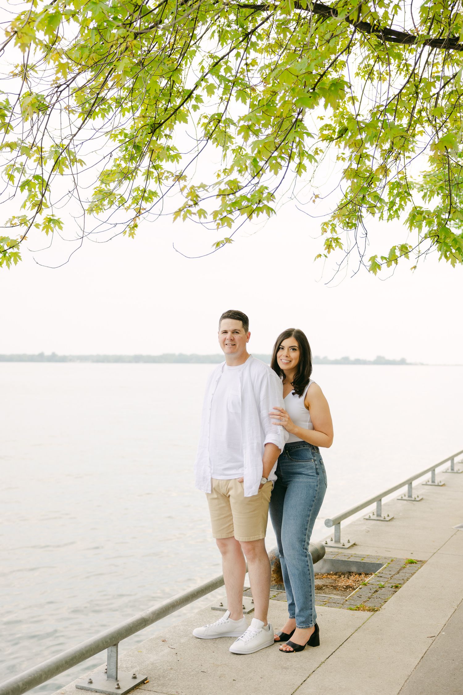 A couple poses together on a waterfront path beneath leafy green branches.