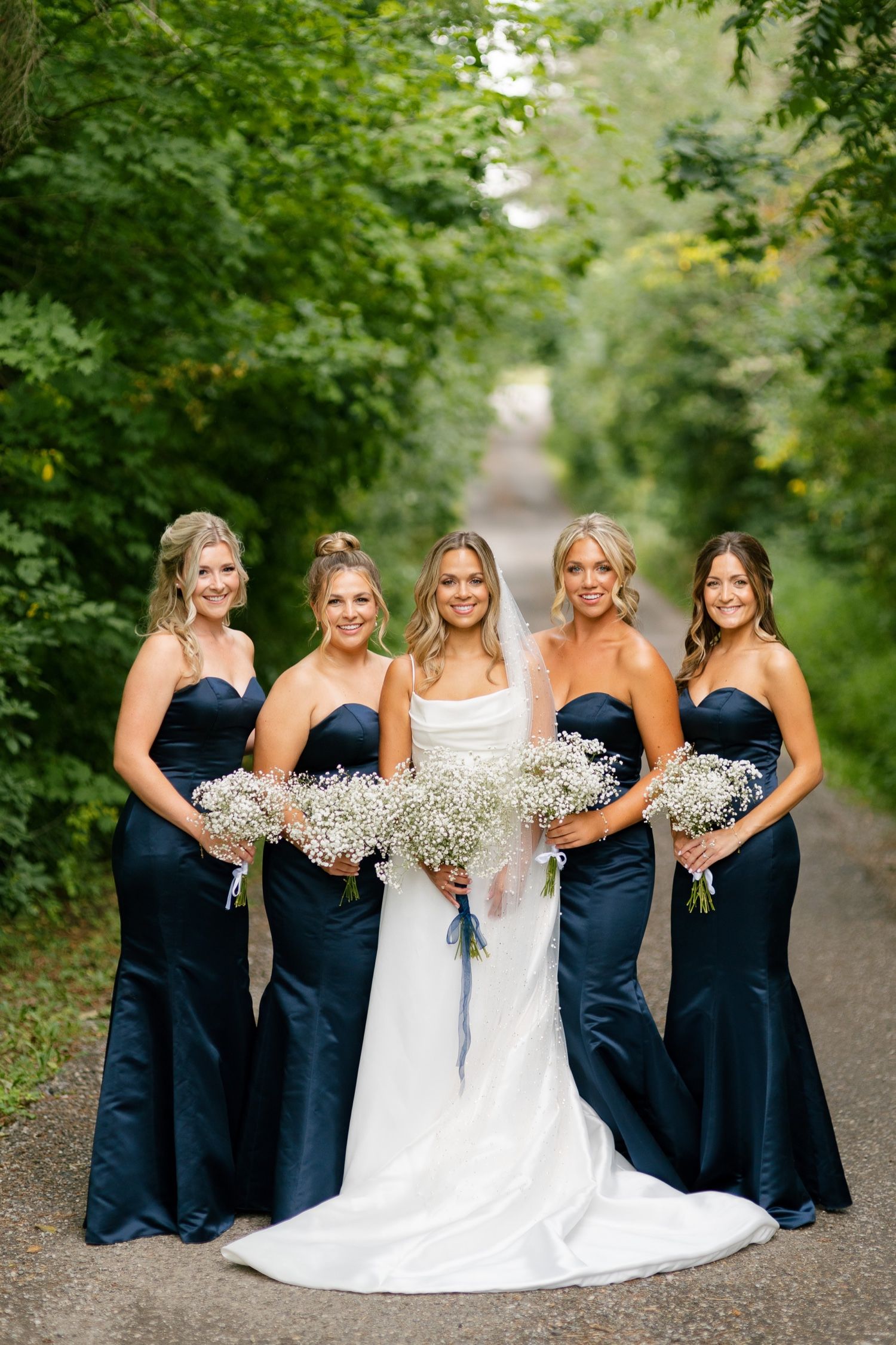 Bridesmaids in navy gowns holding baby's breath bouquets pose with bride on woodland path.