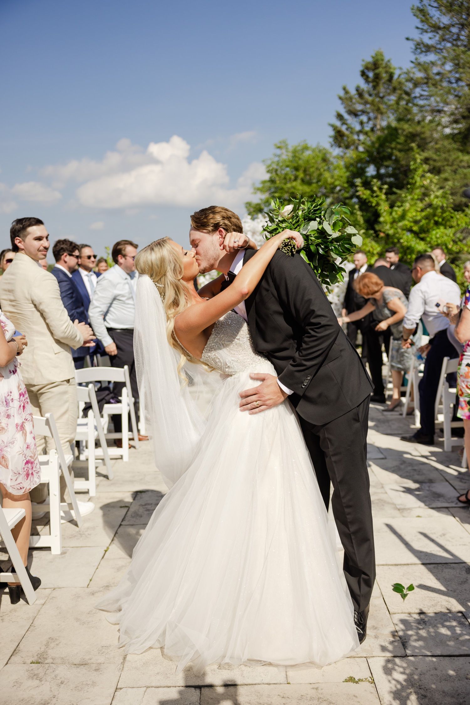 Romantic wedding ceremony kiss under bright blue skies with guests looking on.