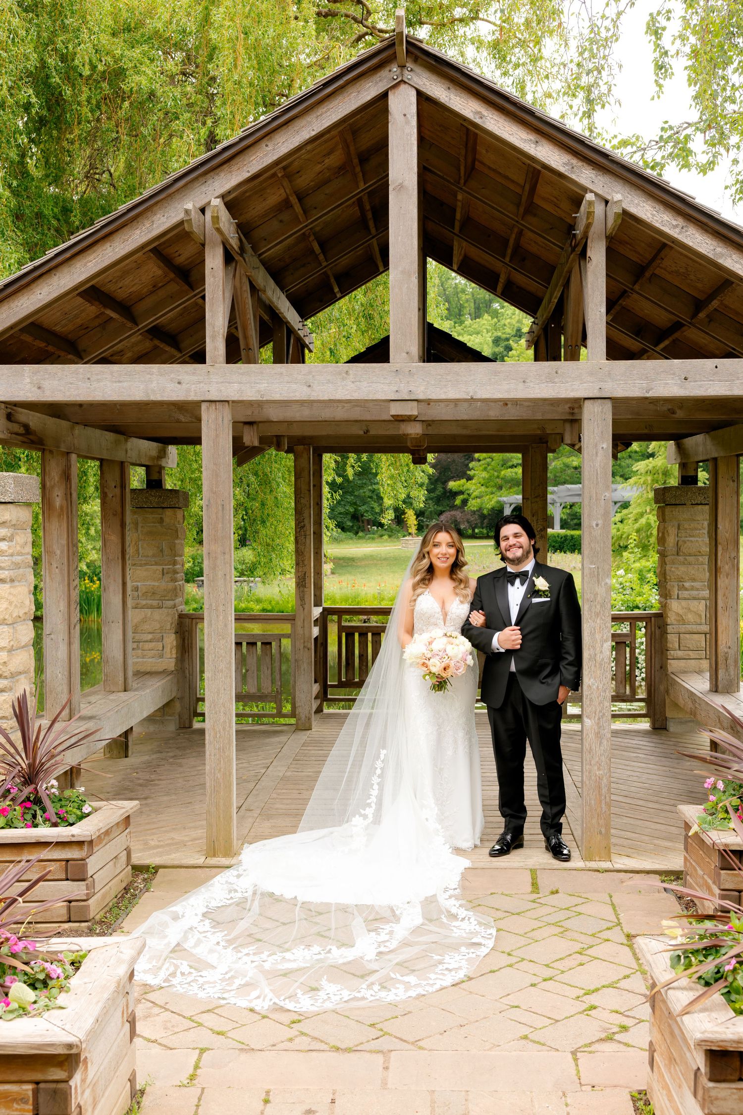 Wedding couple posing under a rustic wooden gazebo surrounded by lush greenery.