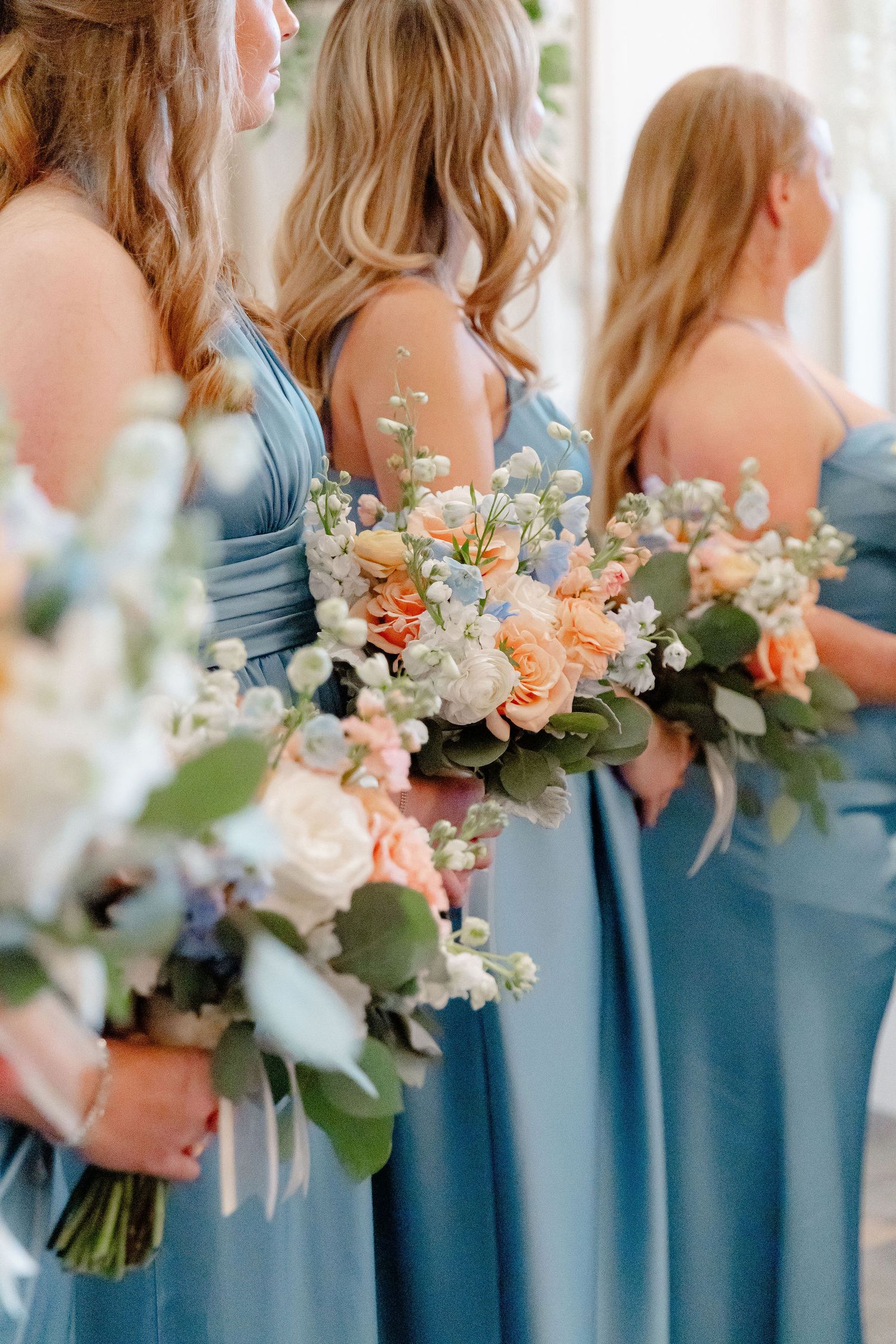 Bridesmaids in dusty blue dresses hold elegant peach and white floral bouquets at a wedding ceremony.