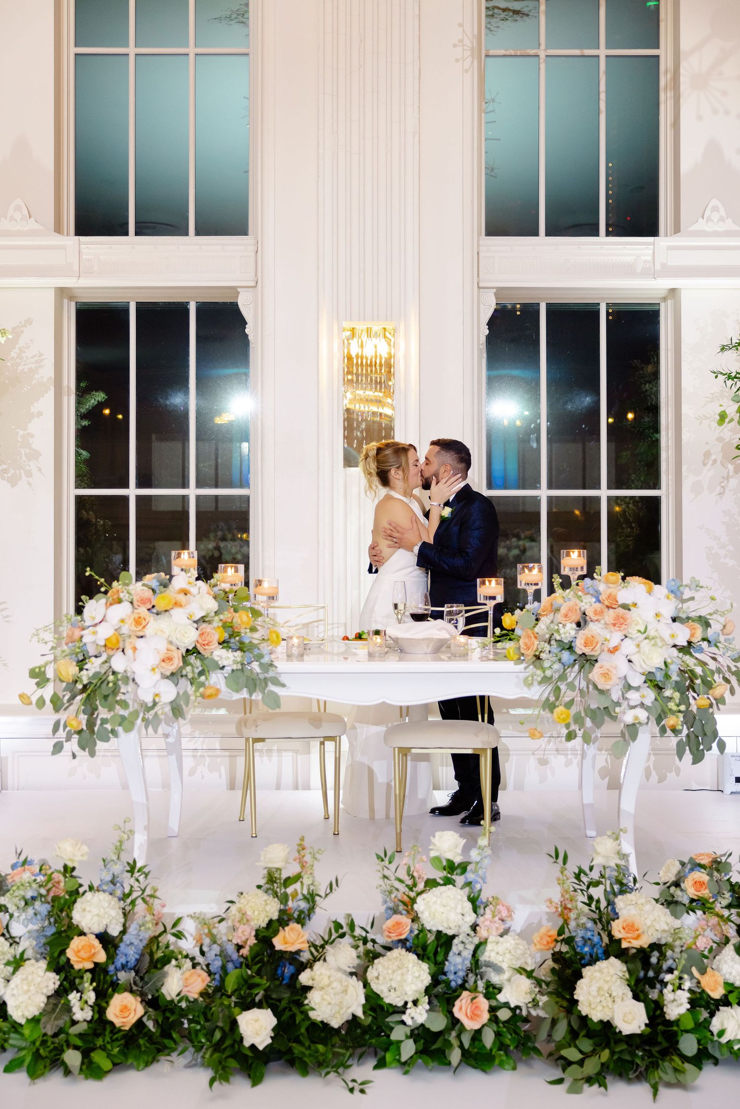 Wedding reception tables adorned with peach and white floral arrangements against large windows.