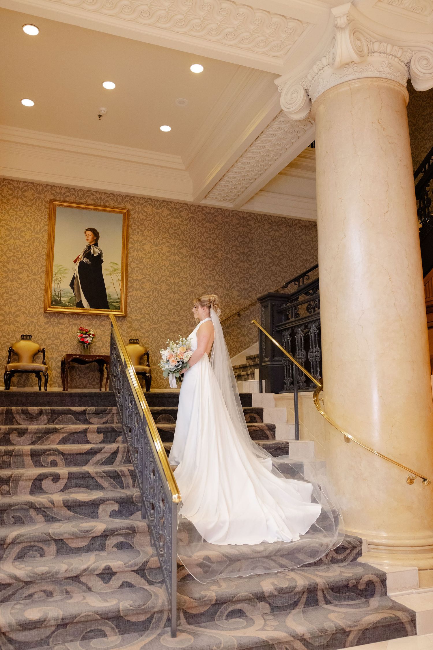 Bride in flowing white gown ascends ornate carpeted stairs with decorative columns and wallpaper.
