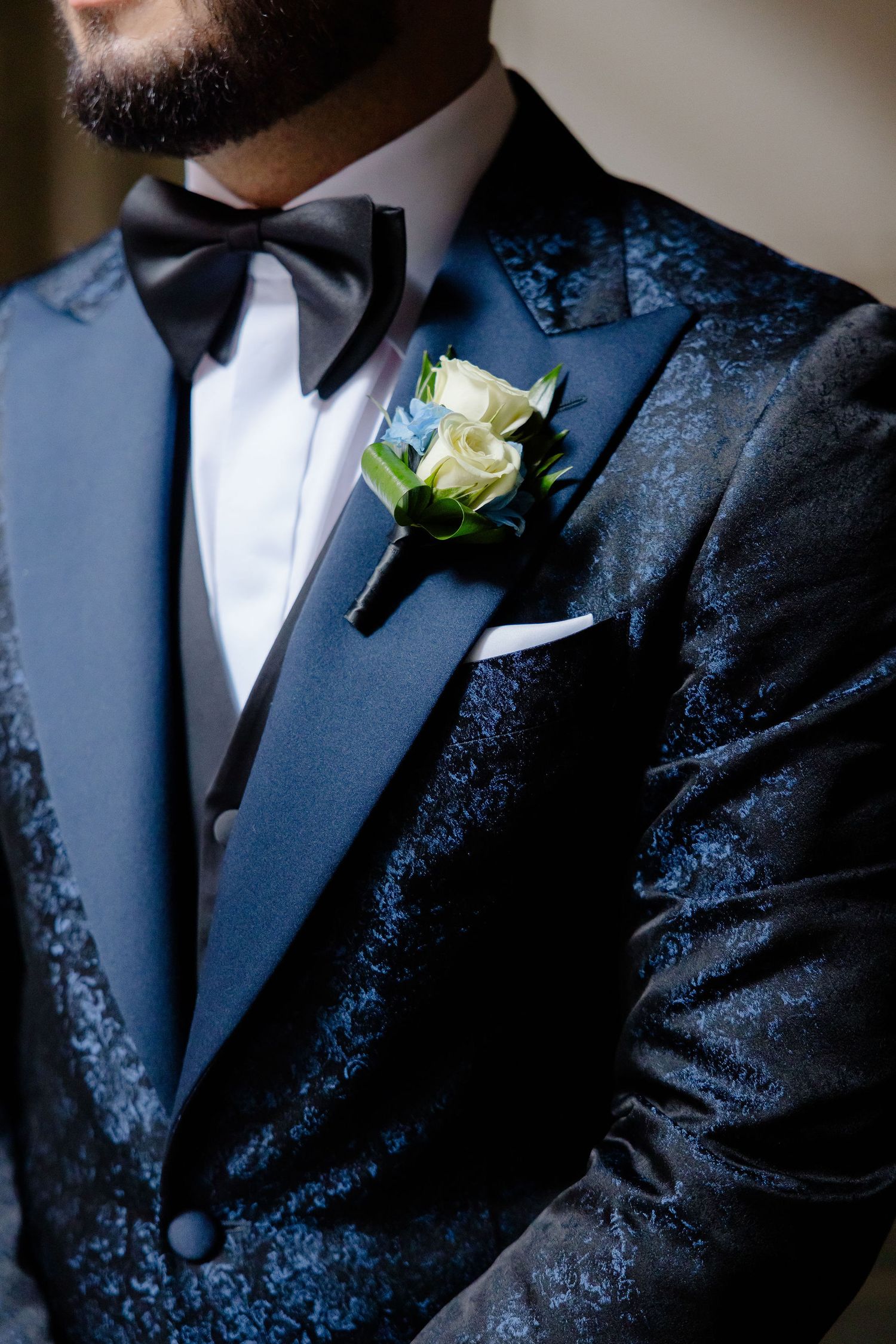 Close-up details of groom's navy jacquard tuxedo with white boutonniere and black bow tie.