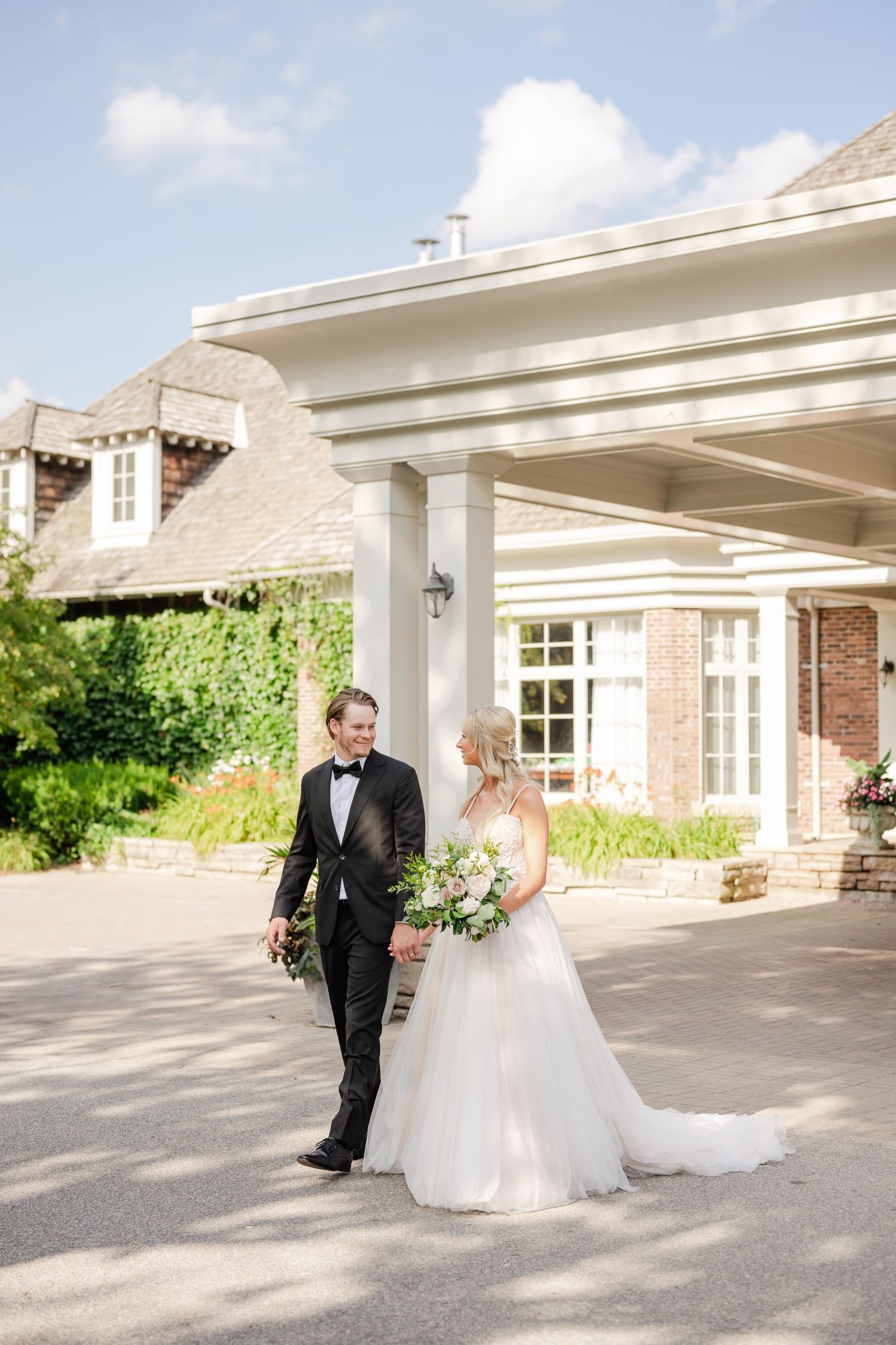 Couple in wedding attire walking under a white columned portico on their special day.