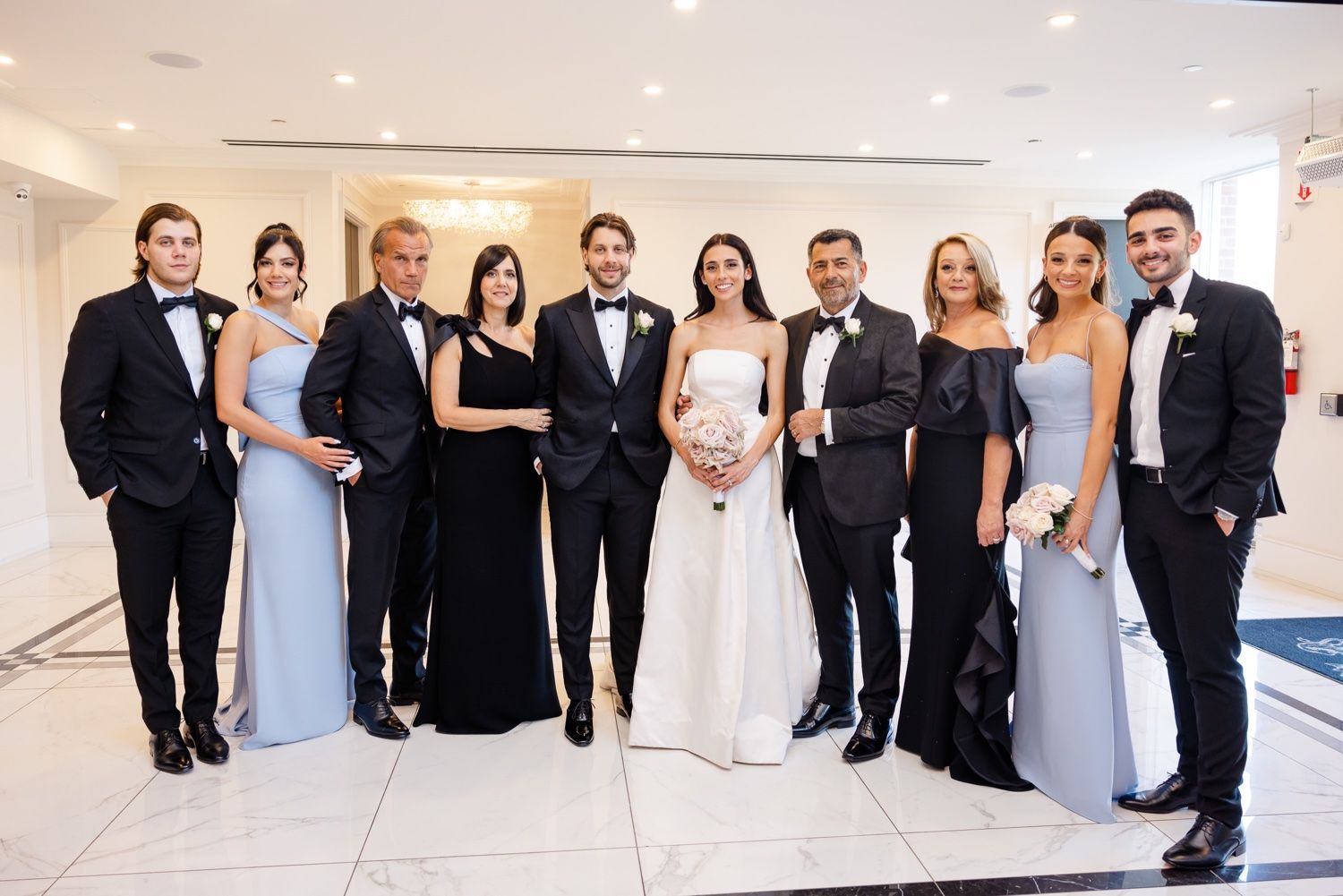 Formal wedding party in black and blue attire poses elegantly in bright indoor setting.