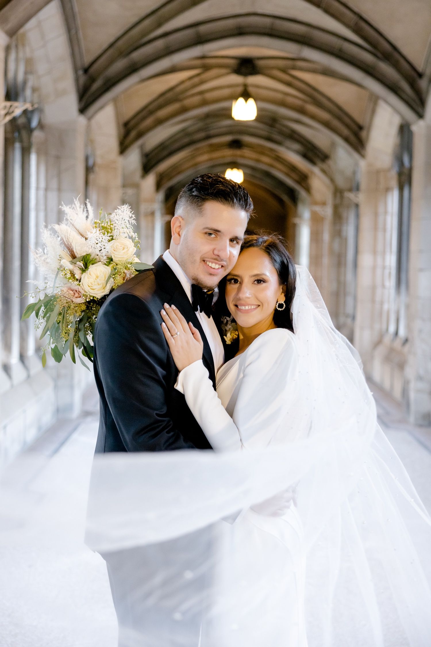 A bright wedding portrait under Gothic arches with flowing veil and white floral bouquet.