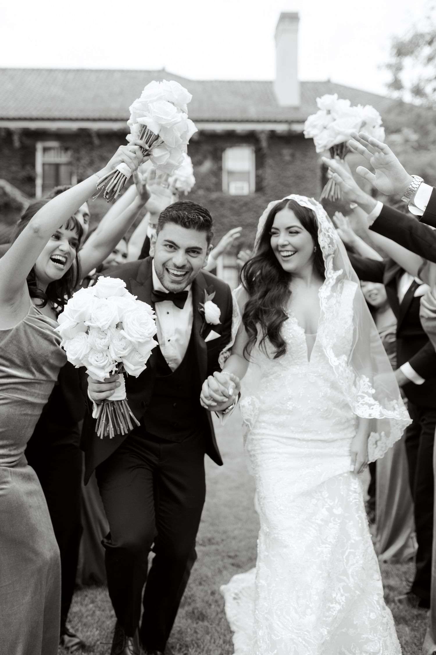 A joyful wedding party raises white bouquets in celebration outside a historic building.