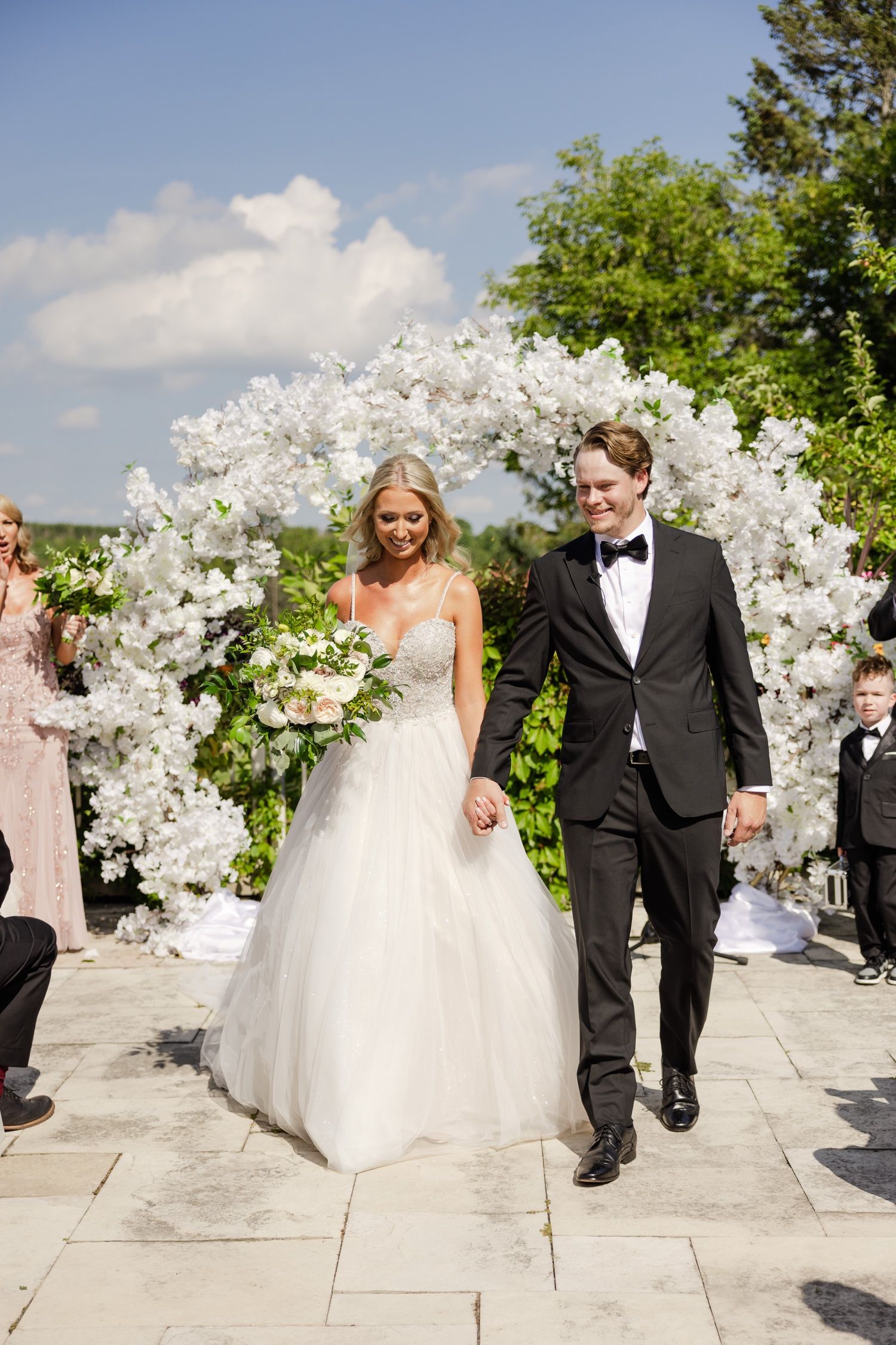Newlyweds walking down the aisle under an elaborate white floral arch during their outdoor ceremony.