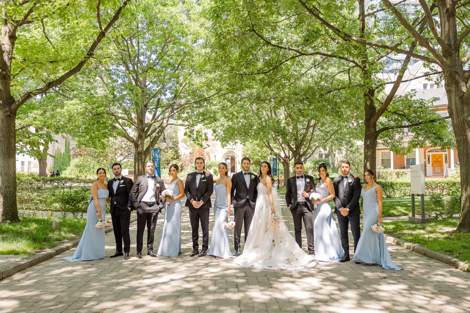 Wedding party stands in formal arrangement under tree-lined pathway.
