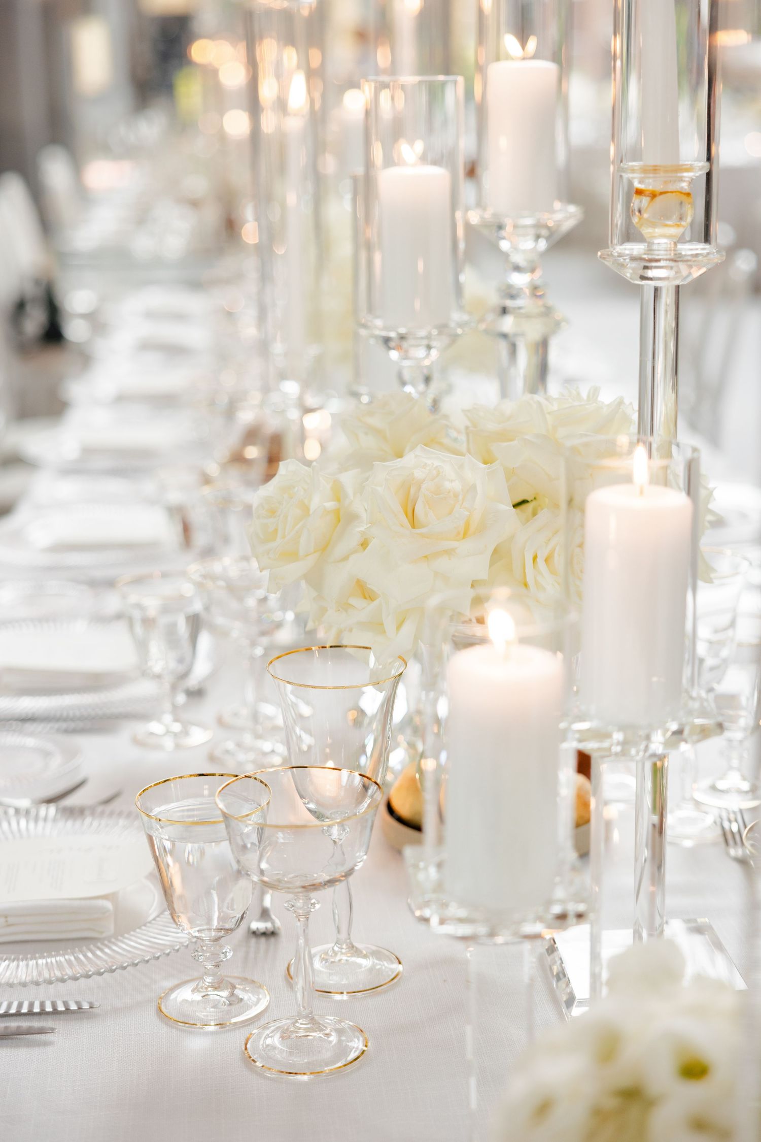Elegant white table setting with crystal glasses, pillar candles in glass holders, and white roses.