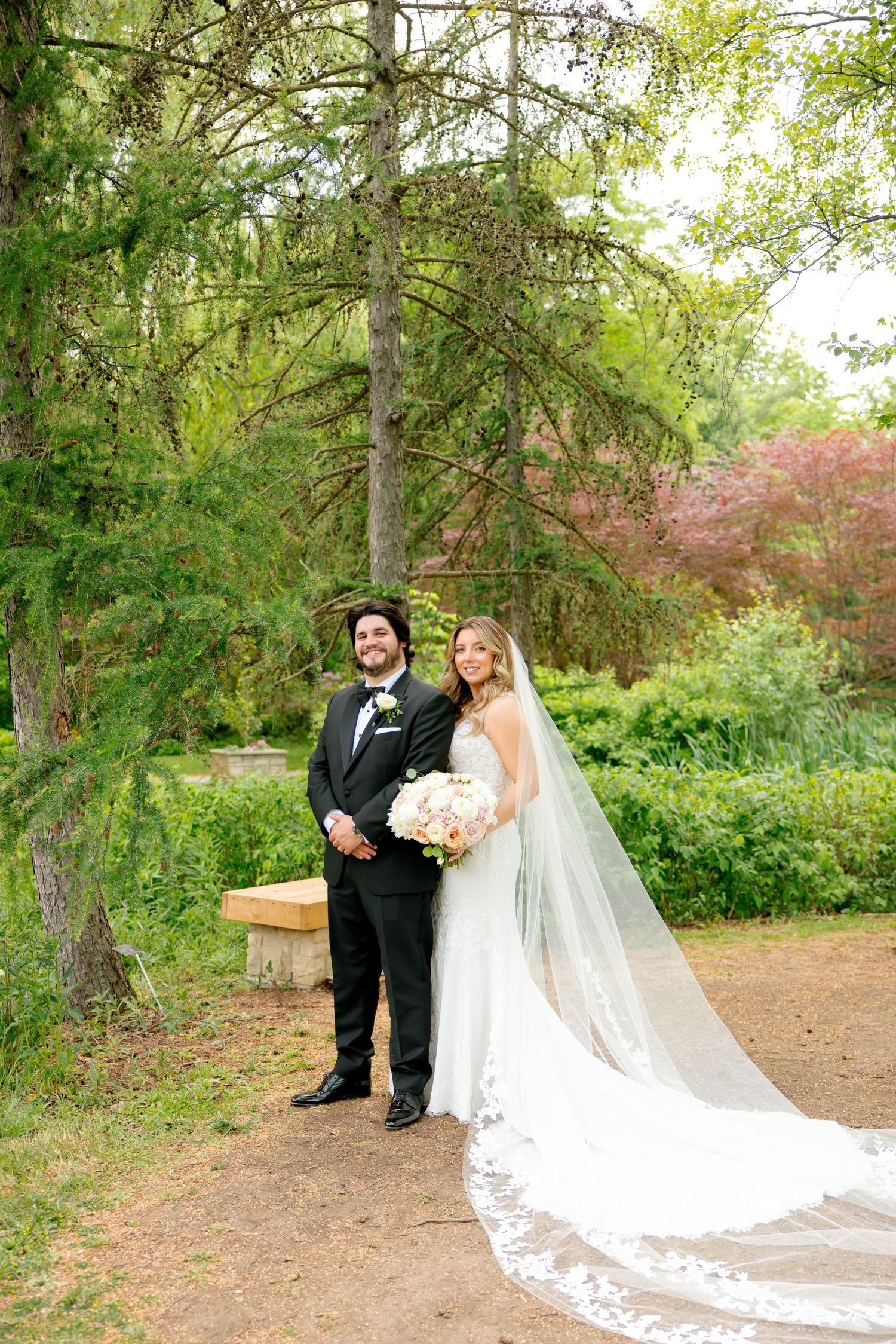 Couple poses for portrait on garden path with dramatic cathedral veil and evergreen trees.