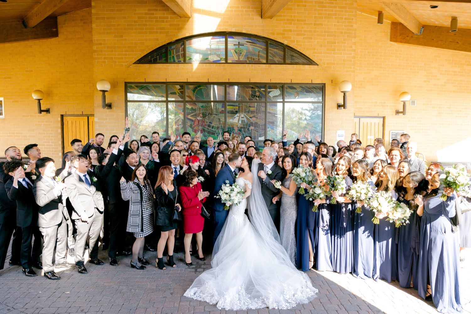 Large wedding party and guests gather outside church entrance for group photo.