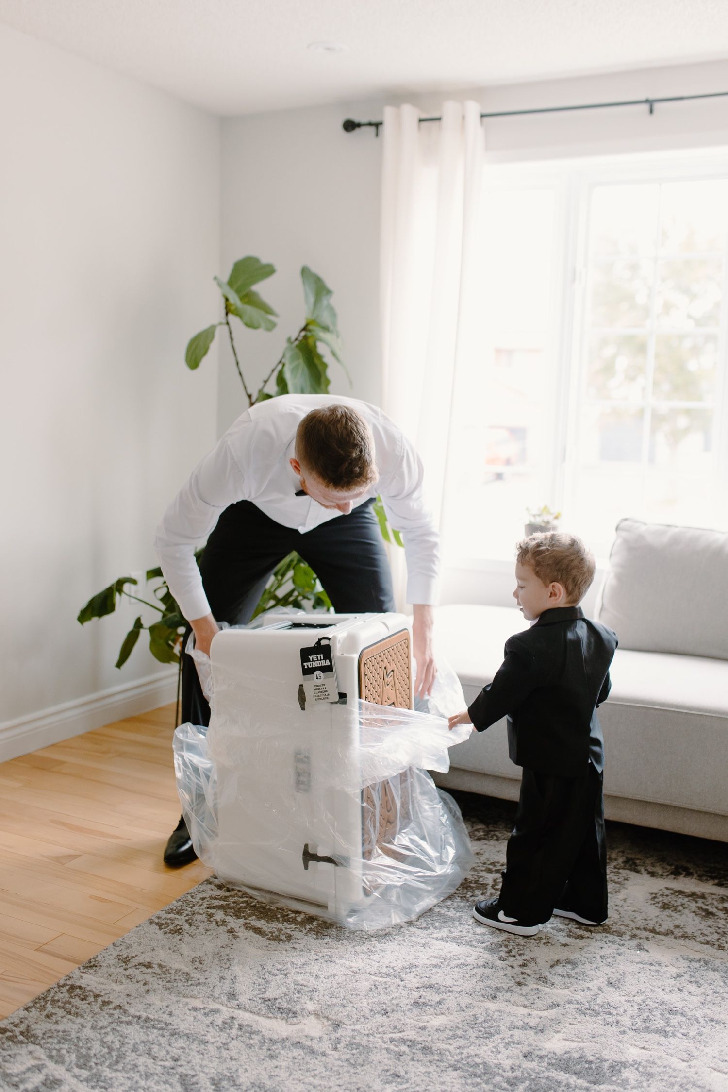 Two figures examine a white box together in a minimalist living room with plants.