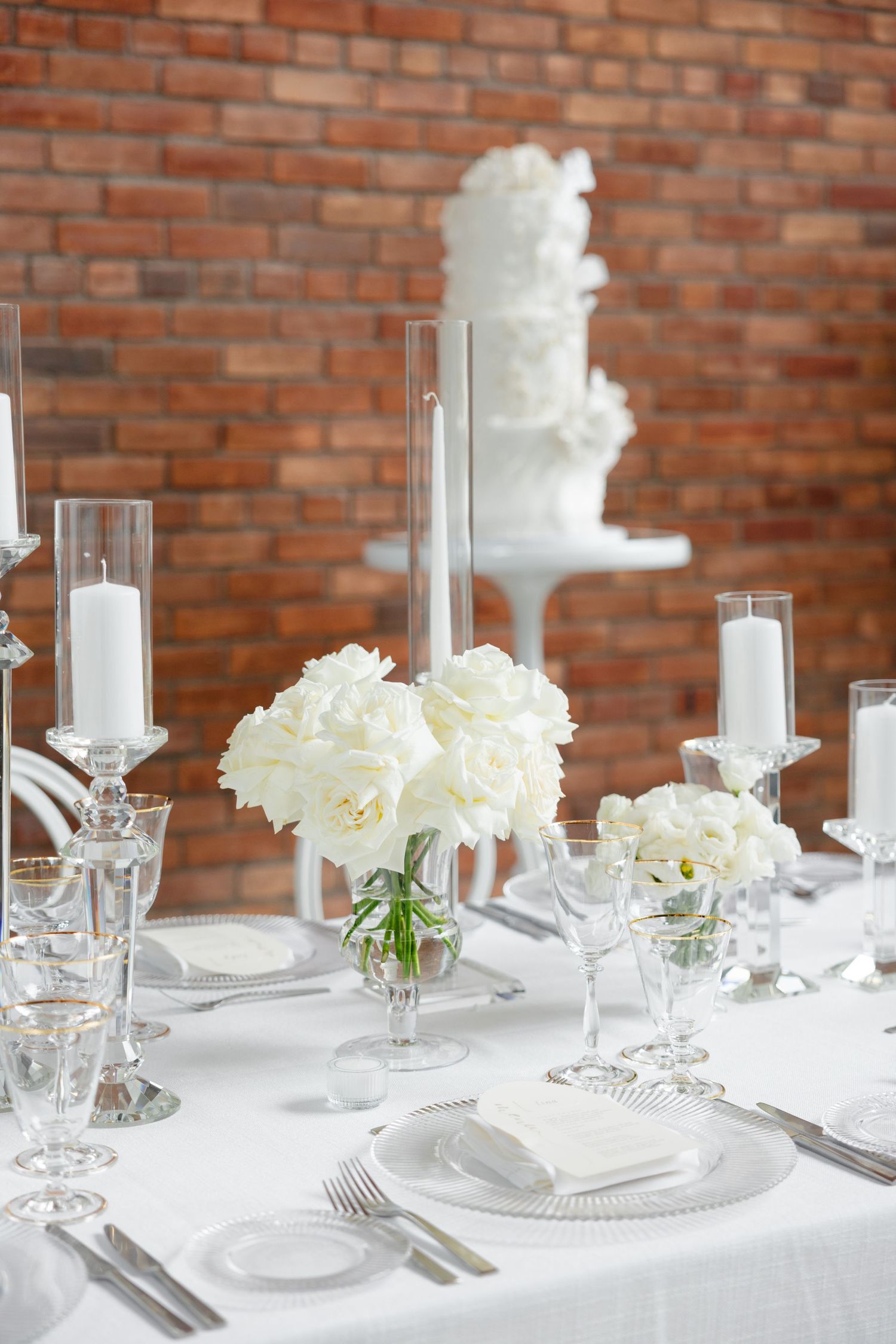 Formal table setting with white roses and tall glass candle holders against a rustic brick backdrop.
