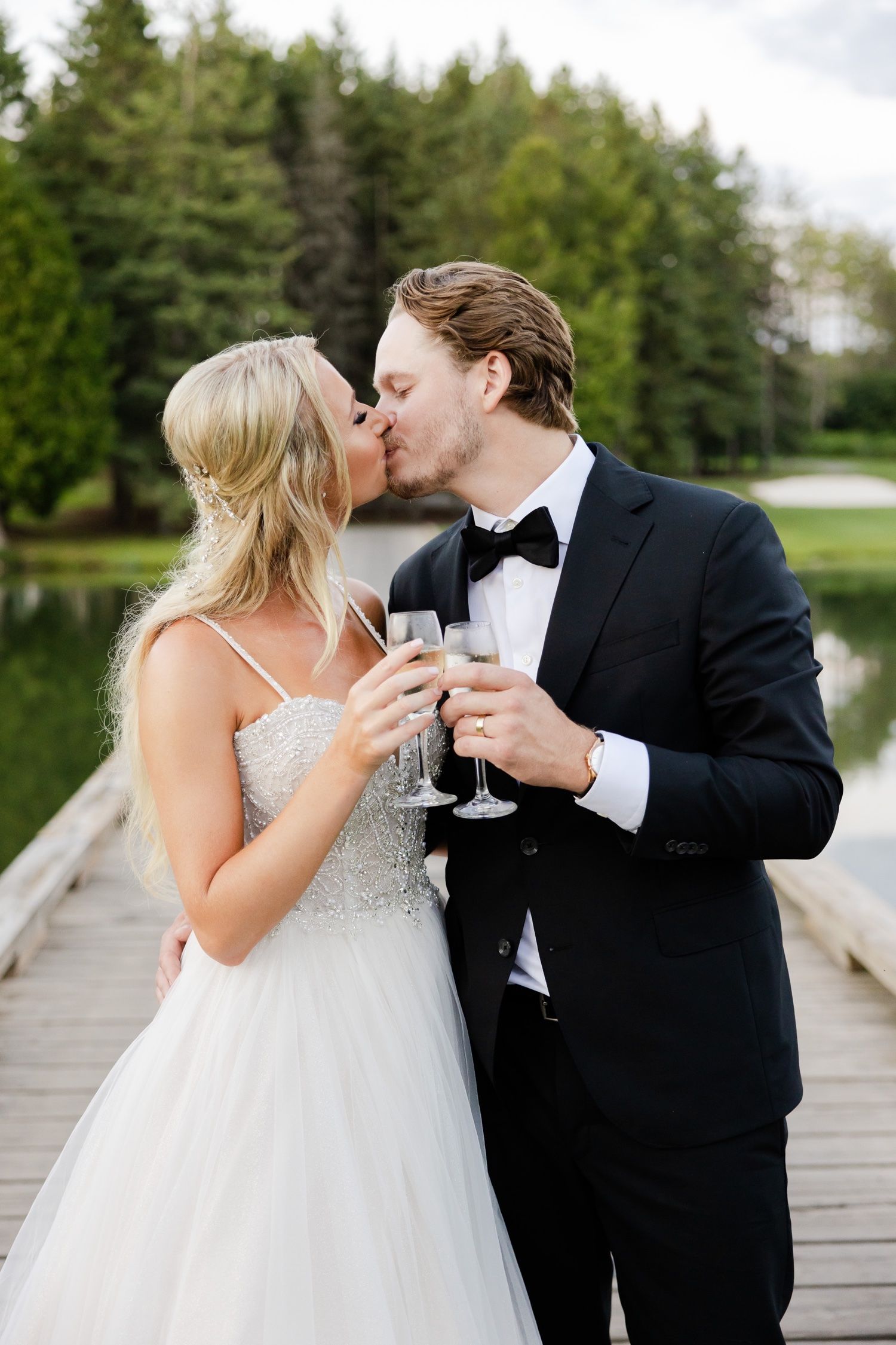 A romantic moment between newlyweds sharing champagne on a wooden dock by a lake.
