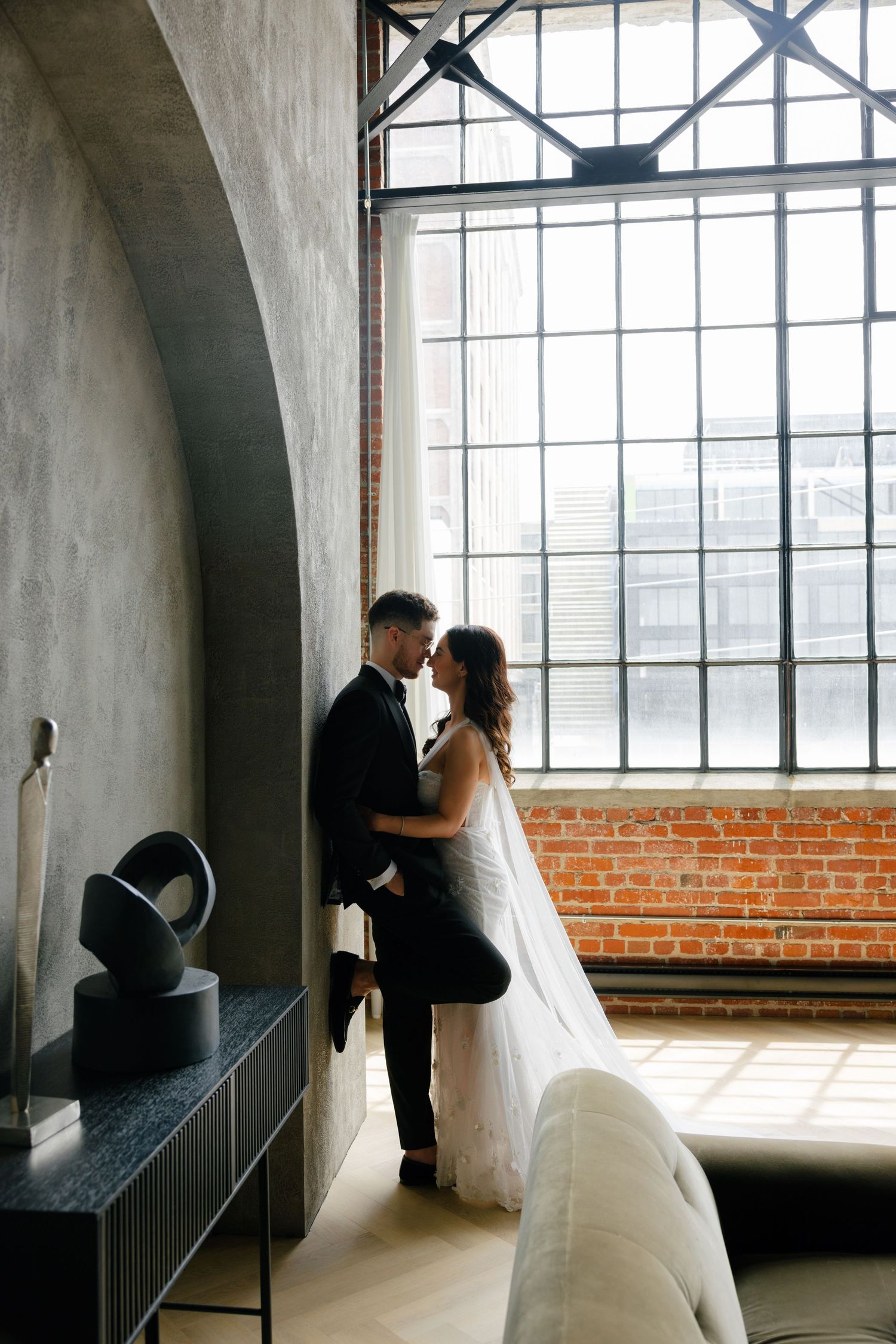 Wedding couple embraces near industrial windows and exposed brick wall.