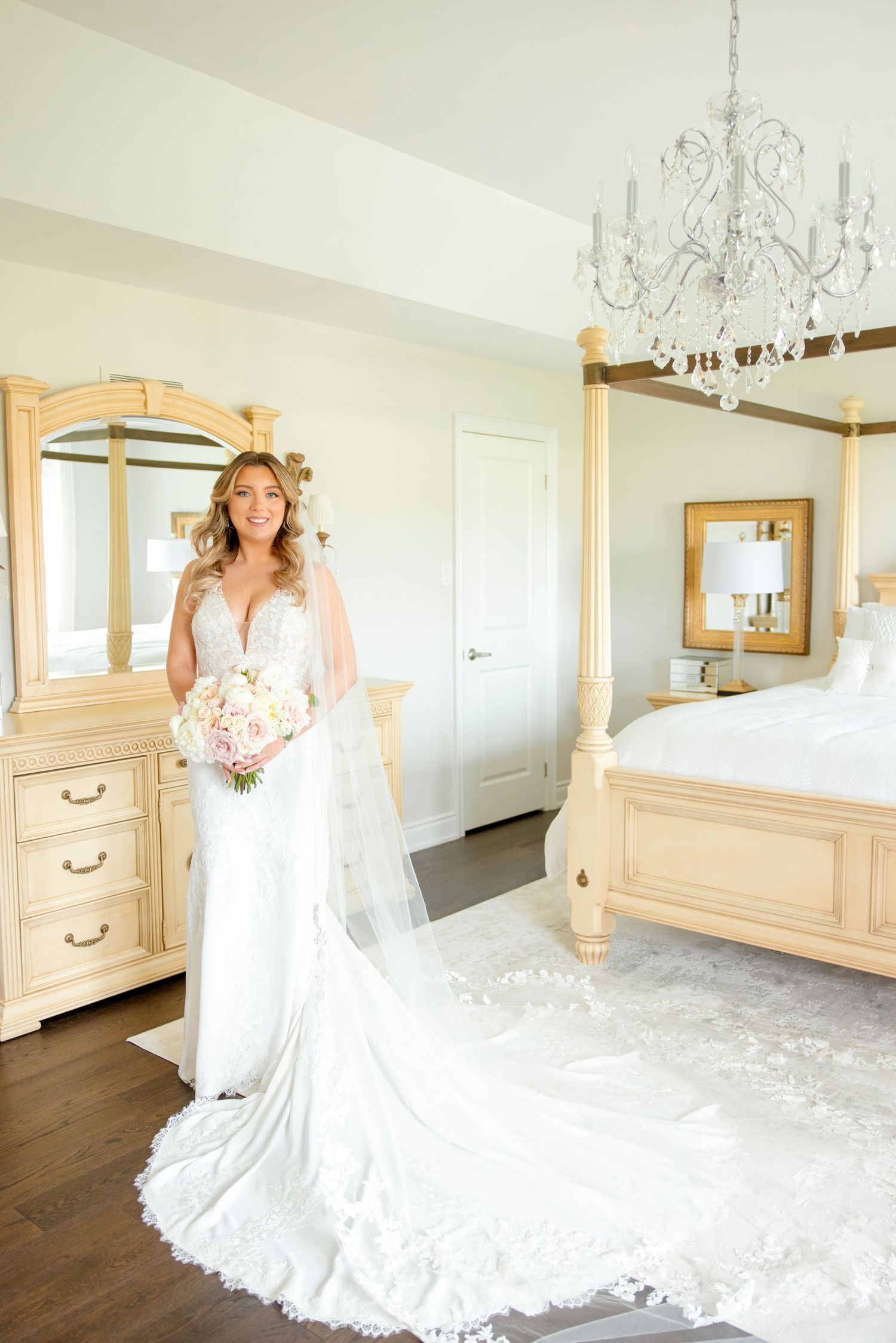 A wedding gown with long train photographed in an elegant bedroom with chandelier and cream-colored furniture.