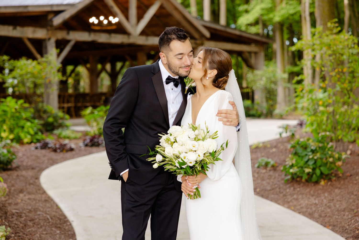 Happy couple embrace on garden path with white floral bouquet and rustic pavilion backdrop.