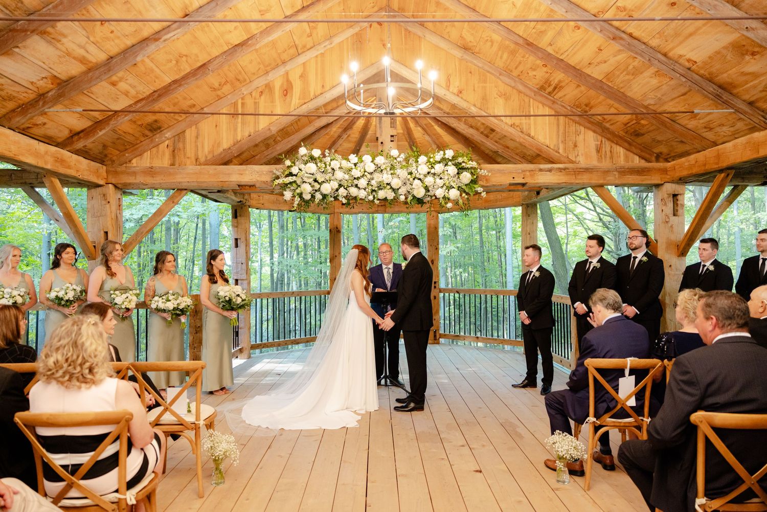 A romantic wedding ceremony in a rustic wooden pavilion with floral decorations and natural forest backdrop.