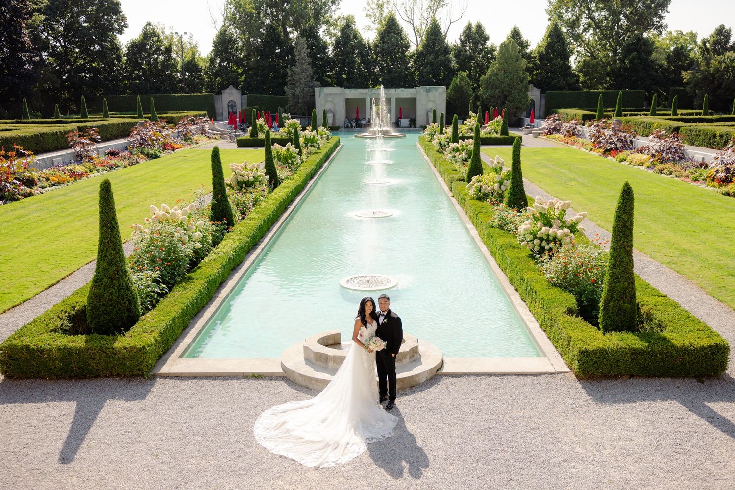 A wedding couple poses by an elegant pool in a formal garden with cypress trees and colorful flower beds.
