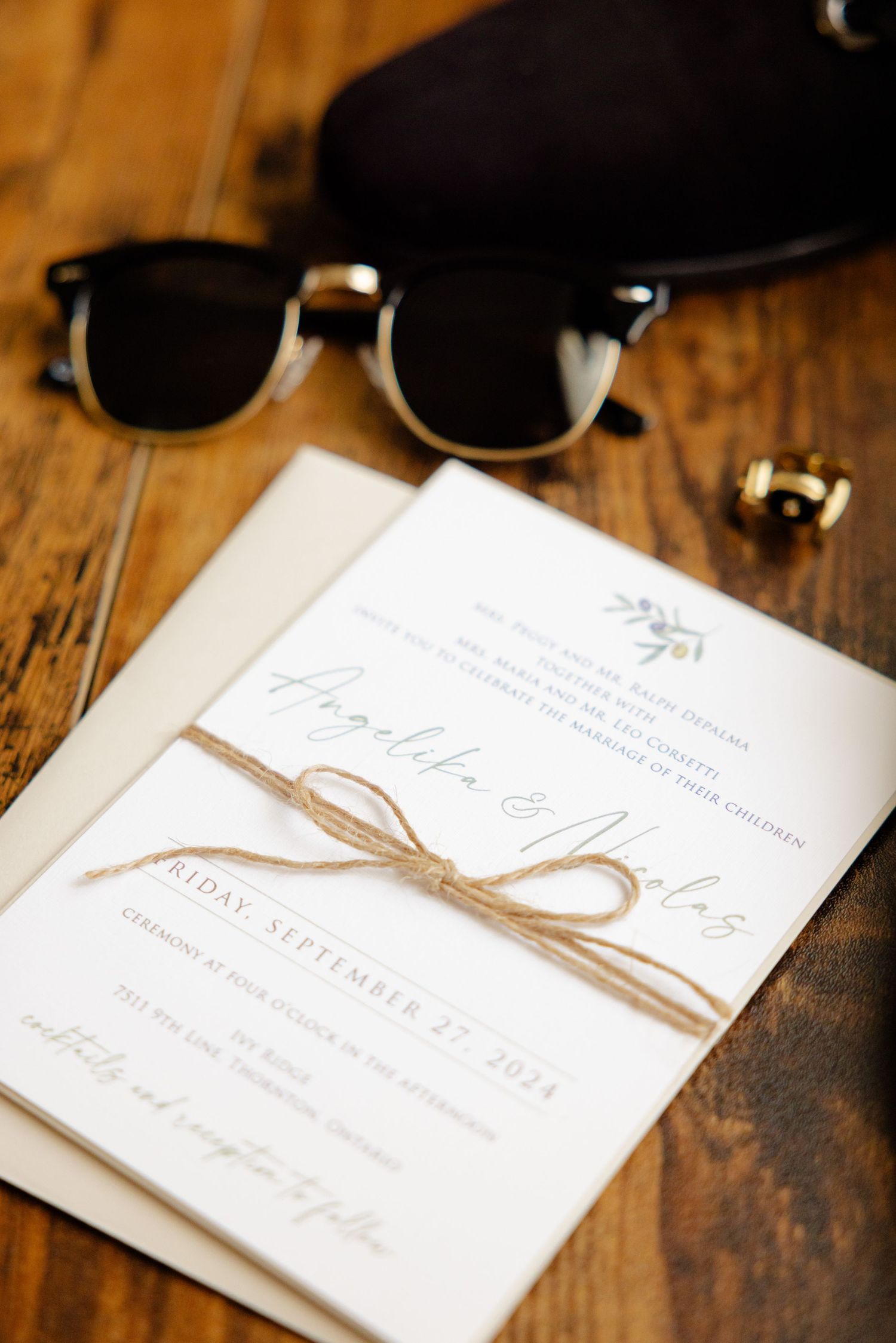 Wedding invitation with twine bow next to sunglasses and ring on wooden surface.