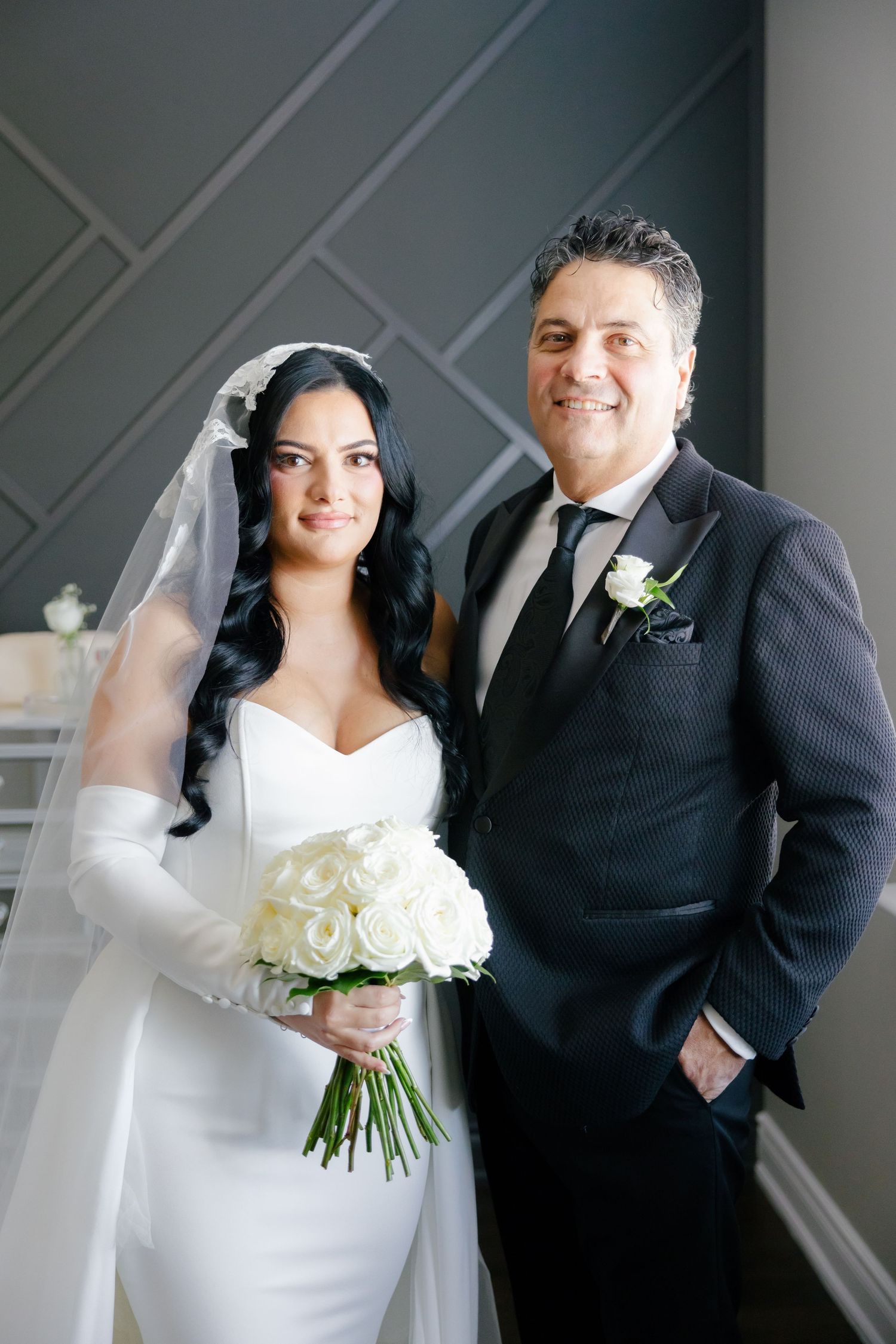 A formal wedding portrait featuring a bride in white with a bouquet posing against a geometric wall backdrop.