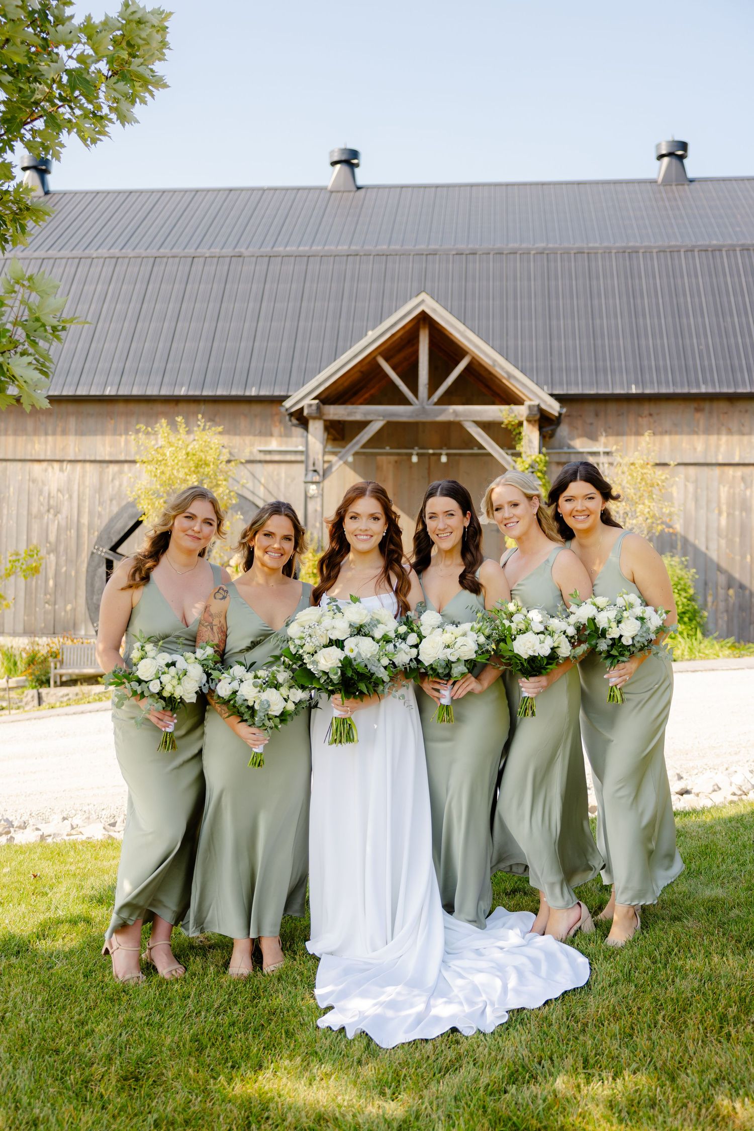 Bridesmaids in sage green dresses pose with bouquets in front of rustic barn wedding venue.