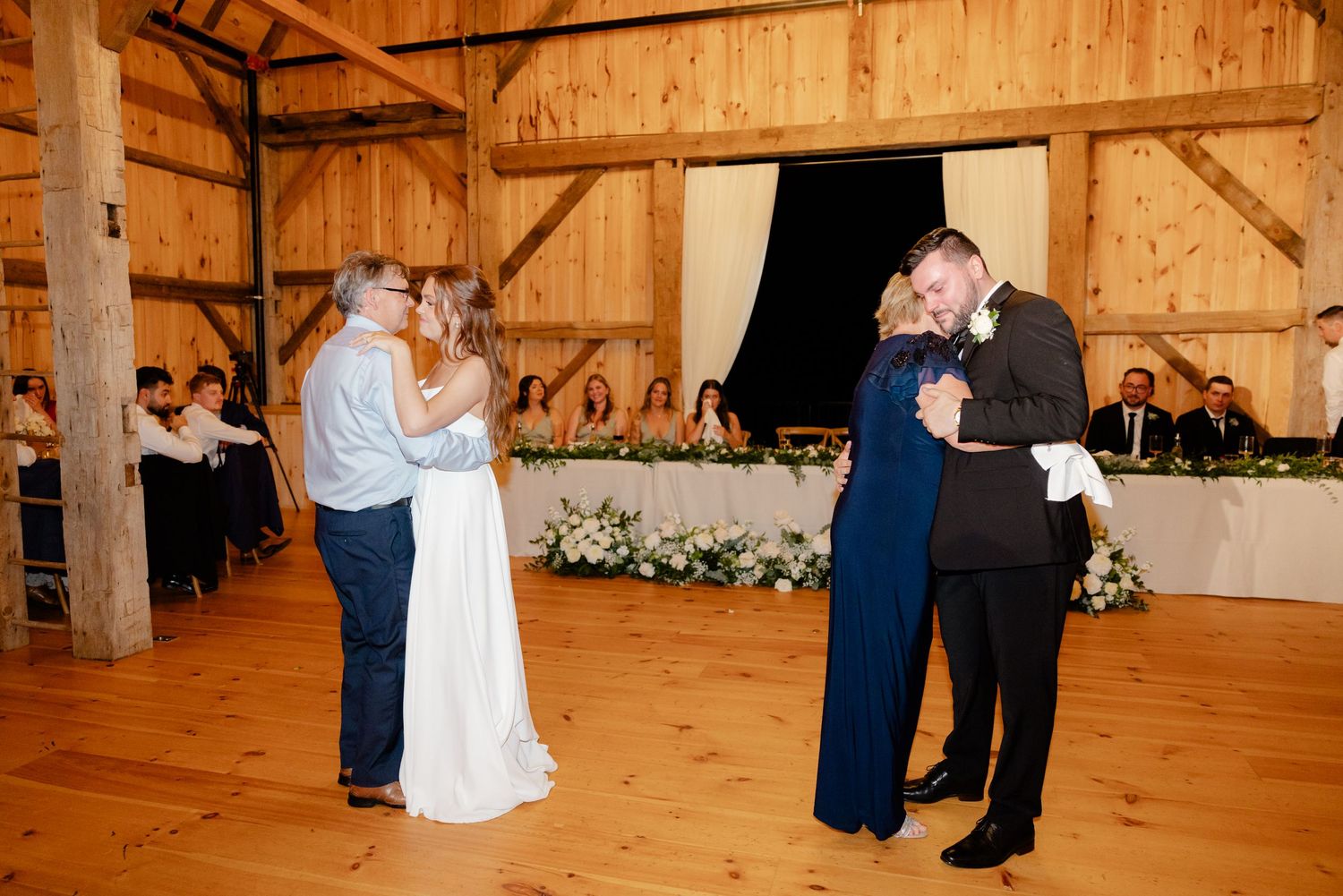 Two couples dance together on a wooden floor during a barn wedding reception.