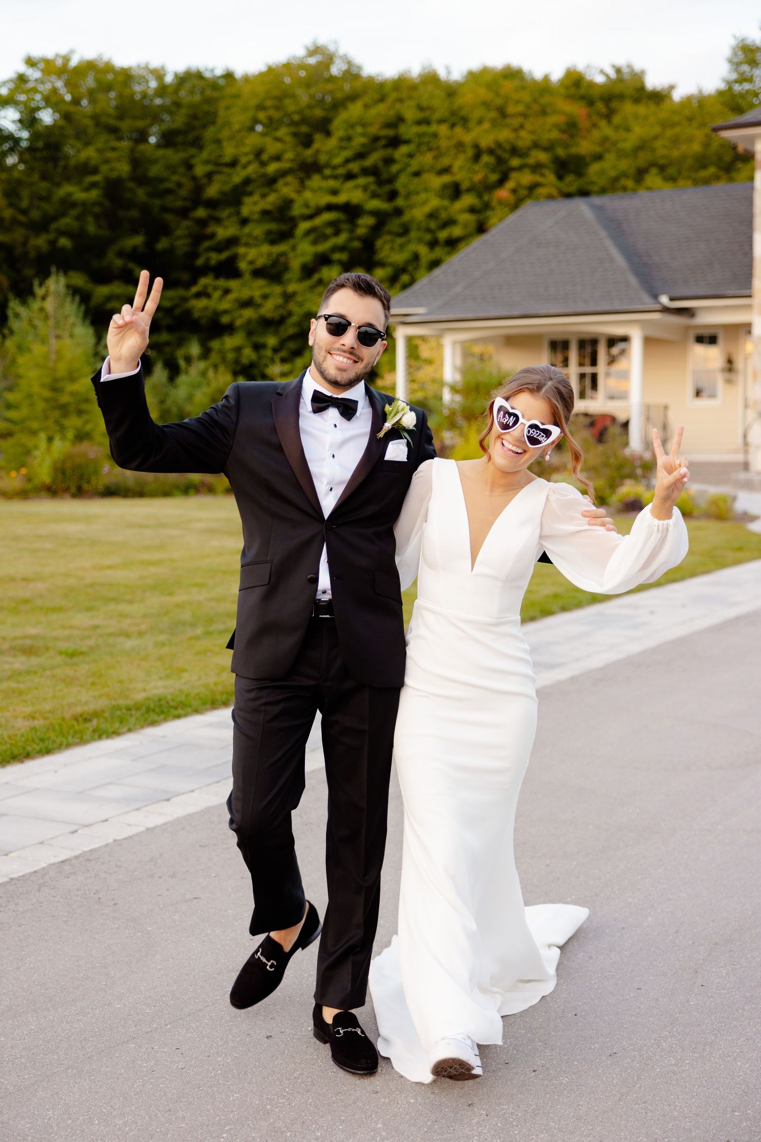 A joyful wedding couple wearing sunglasses makes peace signs while posing on a pathway.