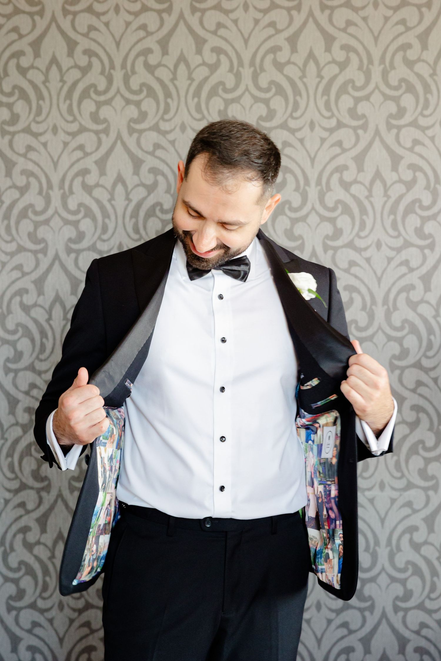 Groom showing off colorful custom jacket lining while smiling against damask wallpaper.