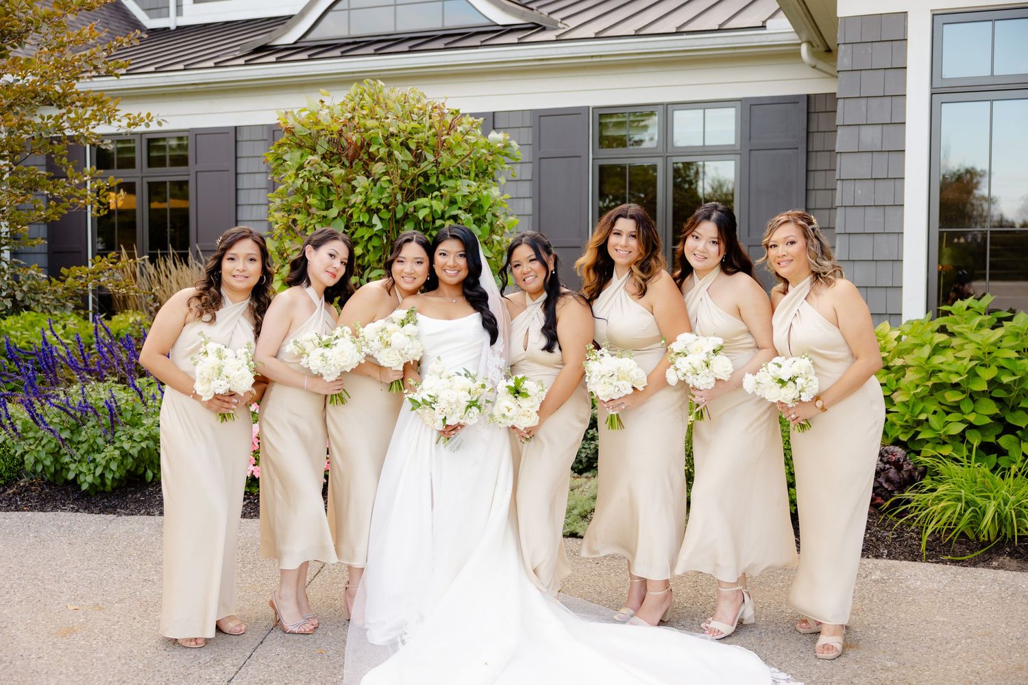 A wedding party in champagne dresses poses in front of a landscaped gray building.