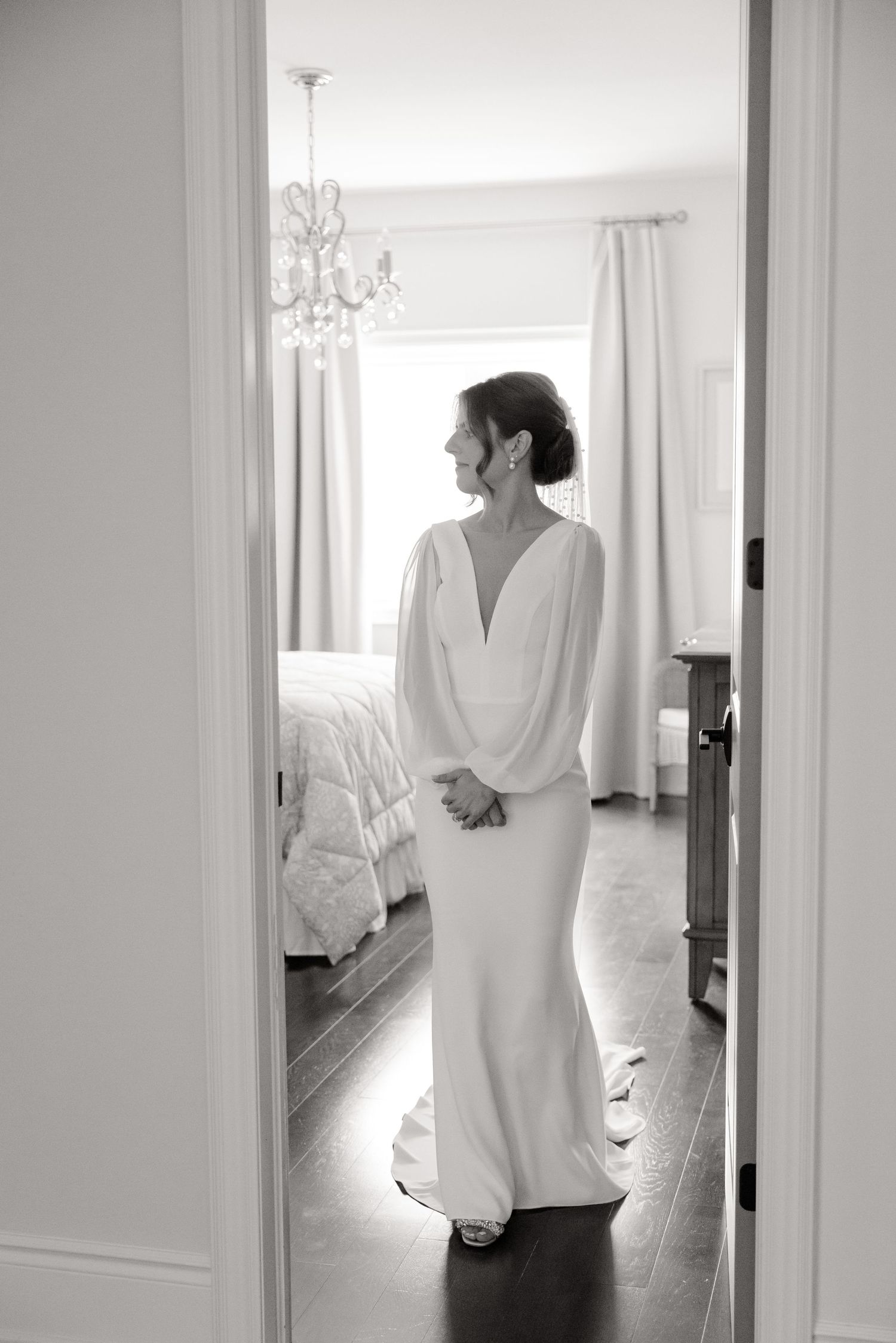 Black and white portrait of bride in flowing white gown standing in bedroom doorframe.
