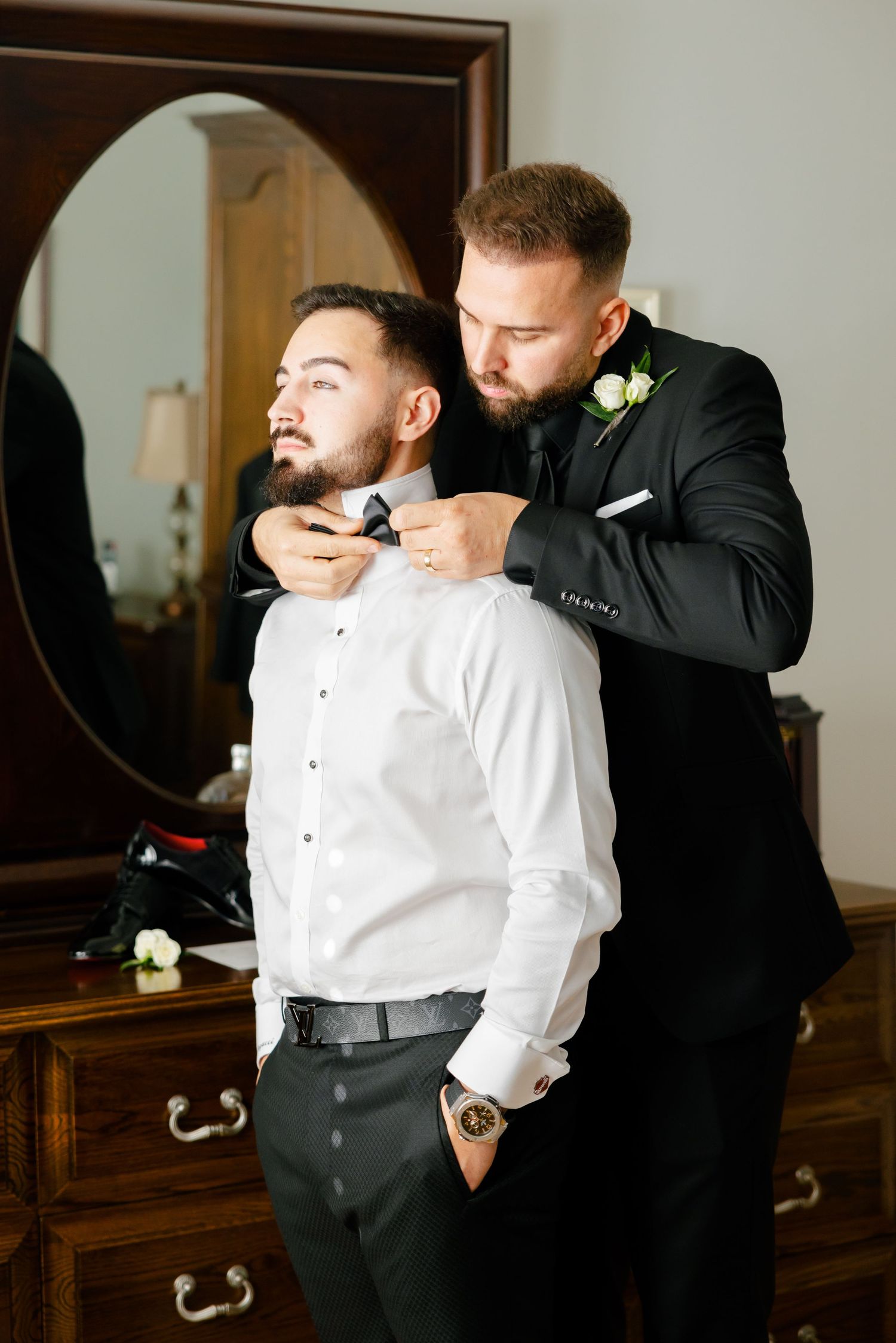 Formal preparation scene with two people adjusting a bowtie near a vintage dresser and mirror.
