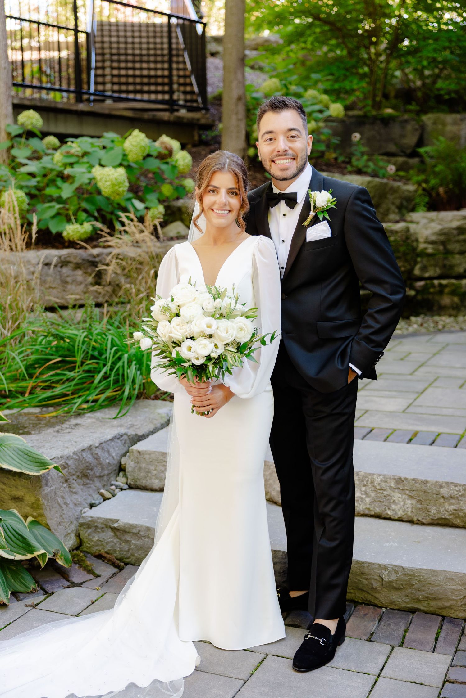 Newlyweds pose together on stone pathway surrounded by lush garden landscaping.