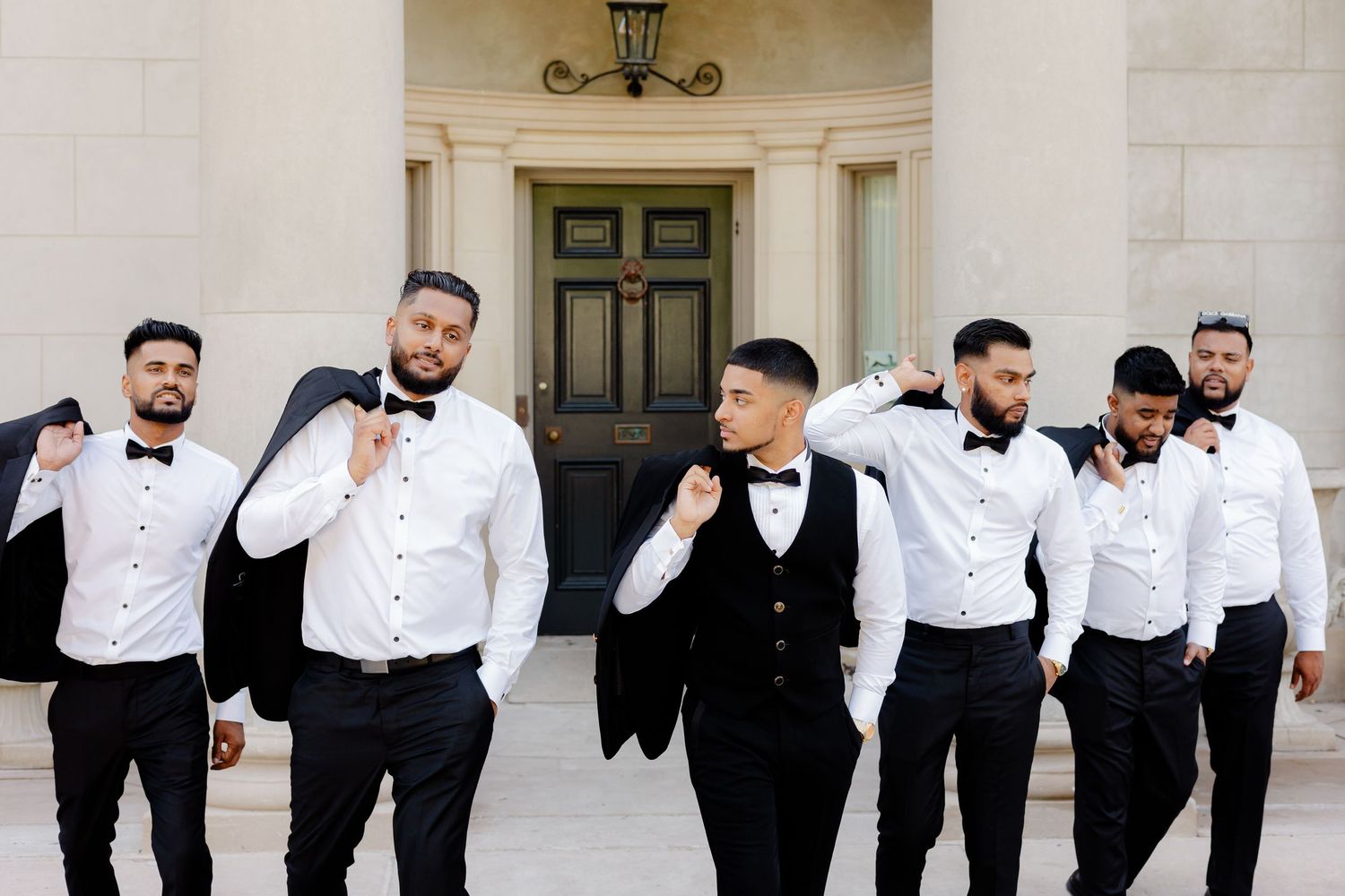 A wedding party of groomsmen in black and white attire pose together on the steps of an elegant venue.