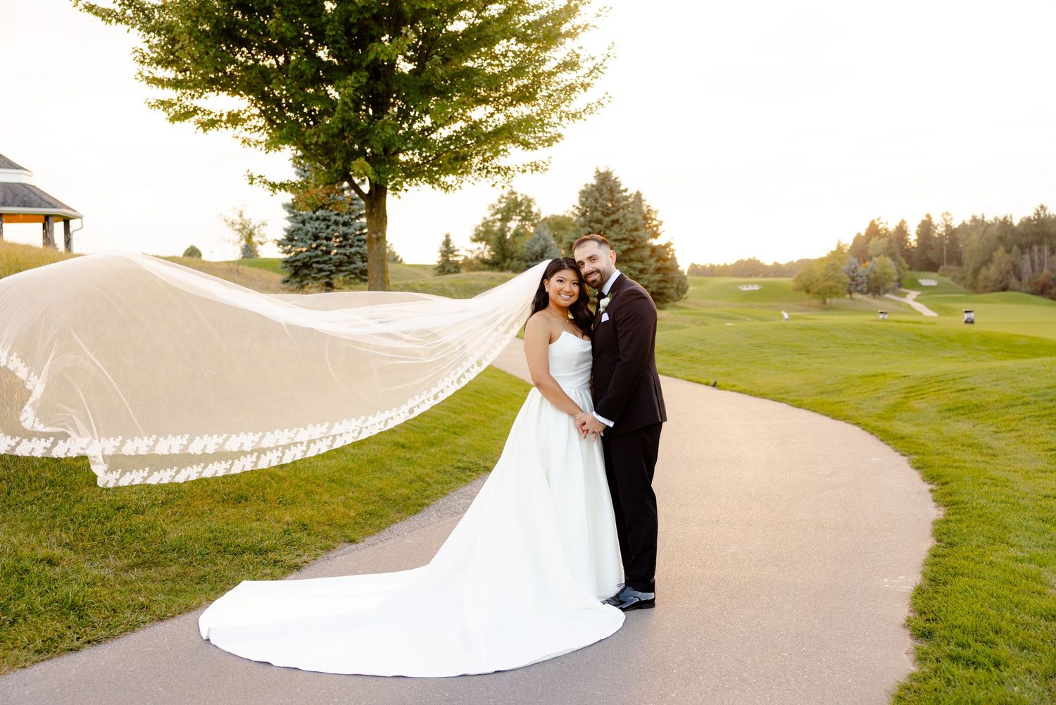 Wedding couple shares an intimate moment as bride's veil flows in the wind on a golf course.