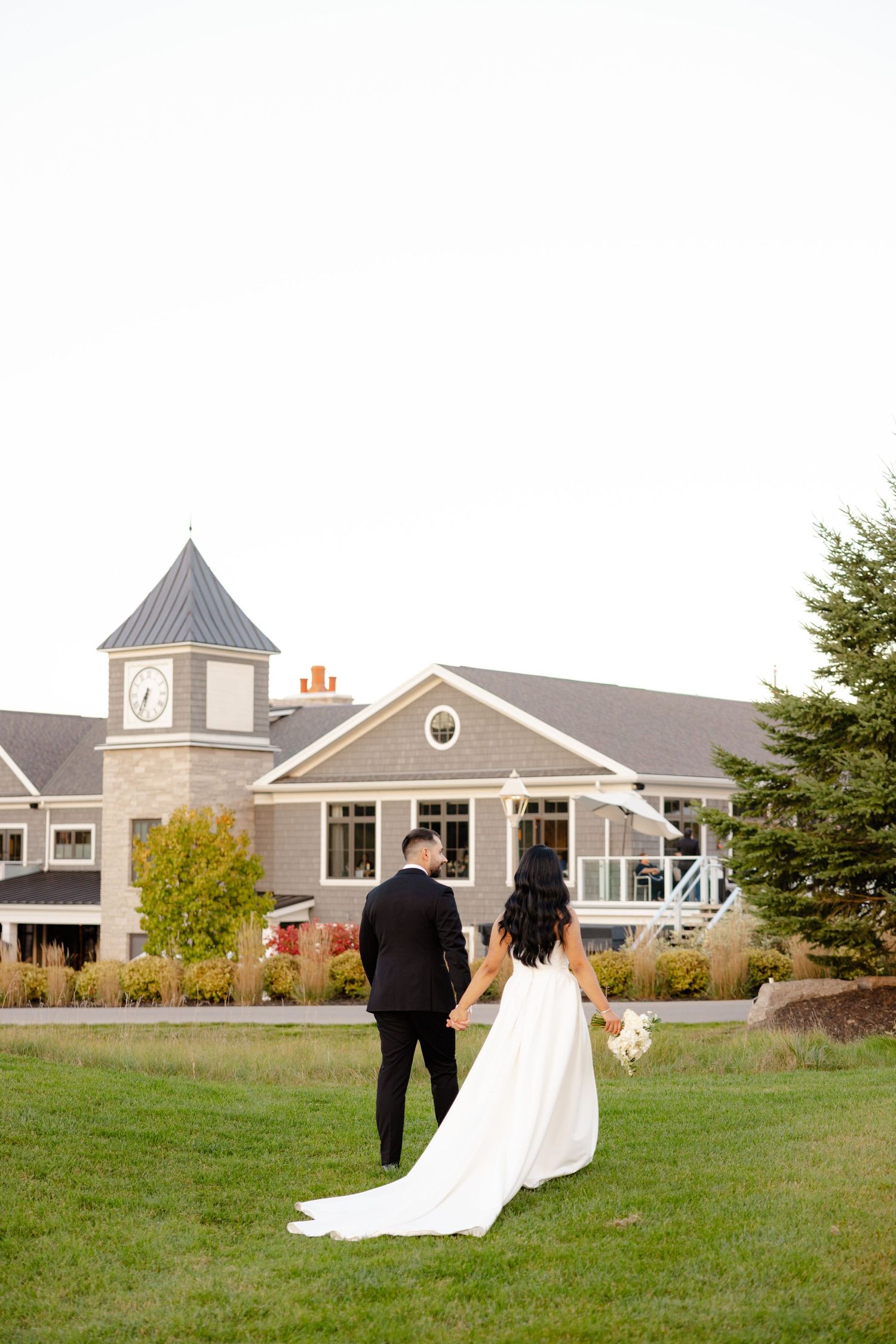 A couple in wedding attire stands on a lawn with a beautiful venue featuring a clock tower behind them.