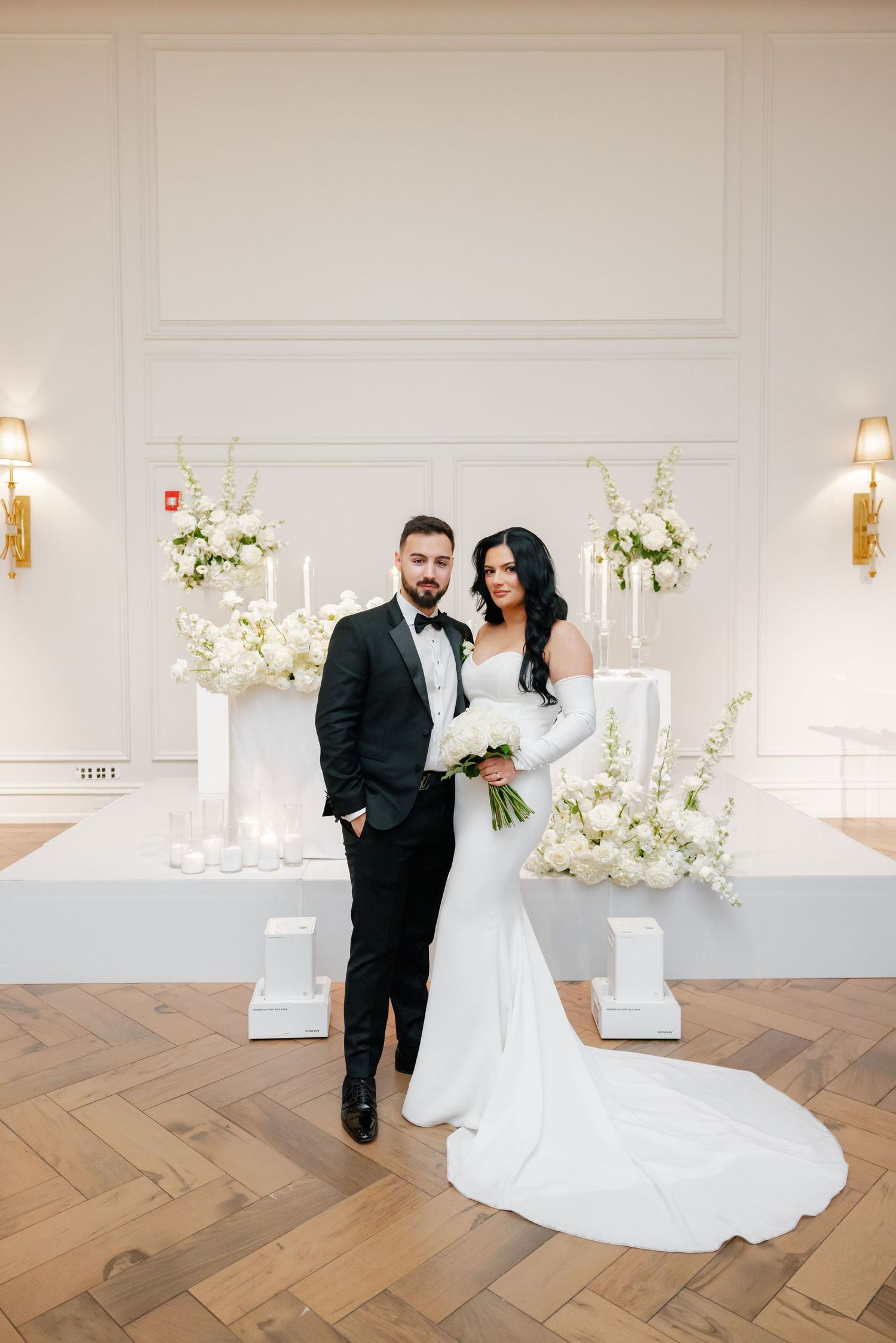 Elegant wedding couple poses in front of white floral arrangements and wall sconces in luxurious venue.
