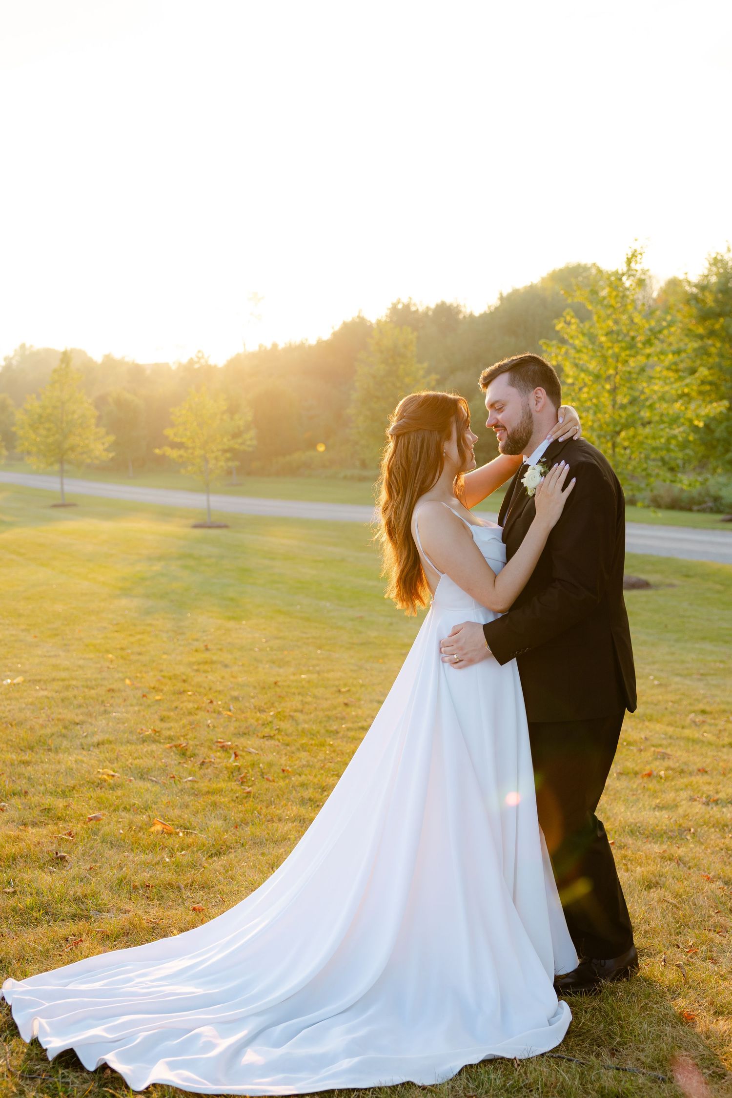 Newlyweds share a romantic moment at sunset in a grassy field, her elegant white gown trailing behind them.