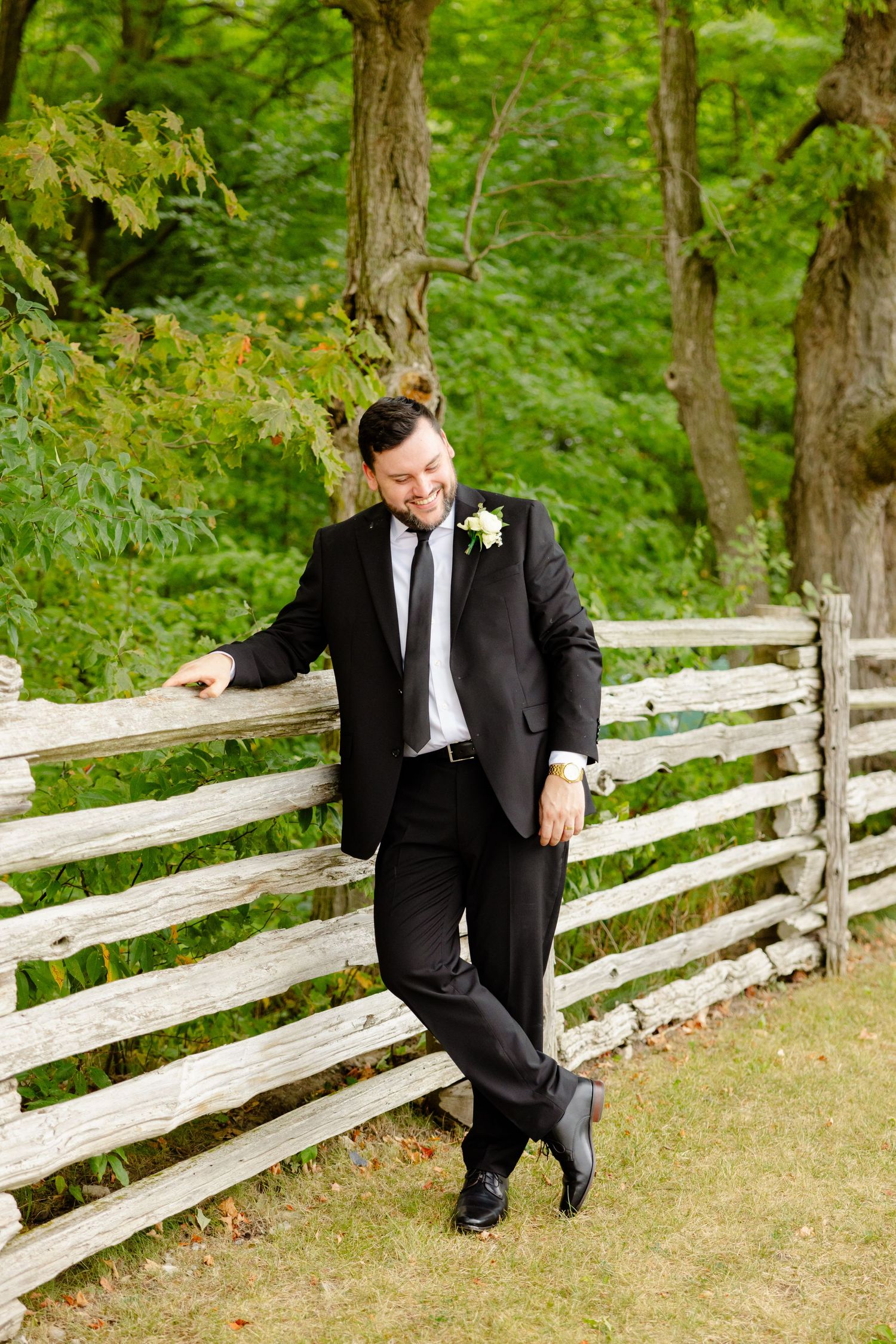 Stylish groom poses casually against wooden fence in outdoor setting.