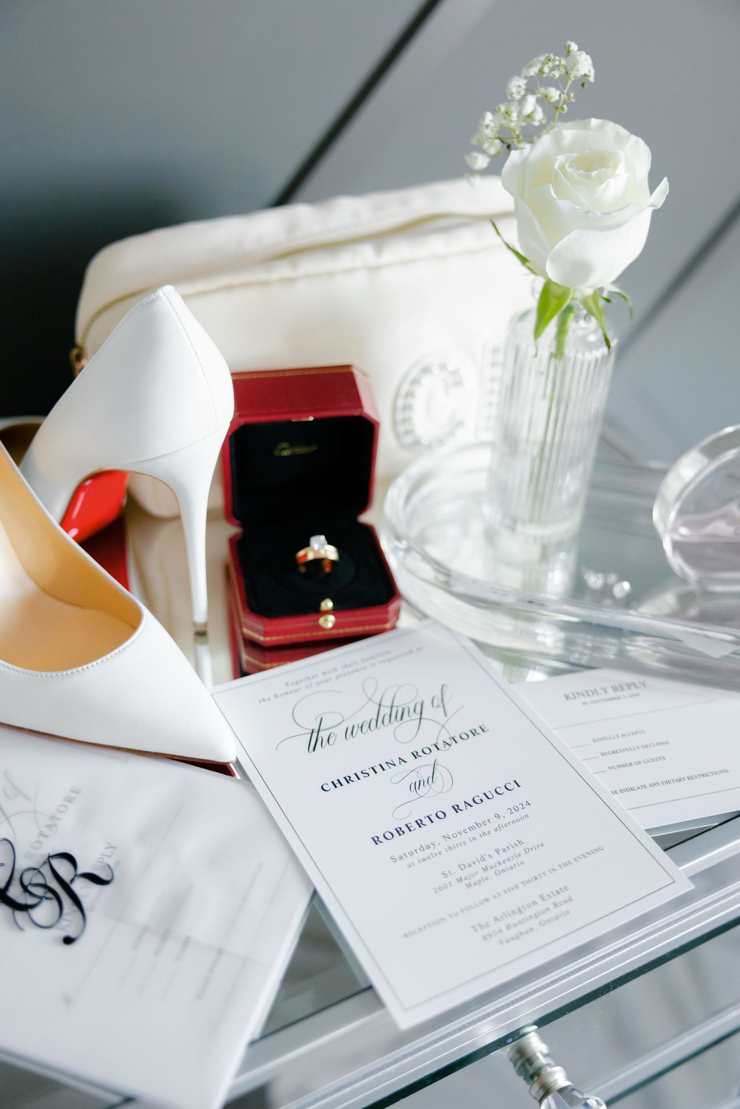 Wedding invitation, white heels, and engagement ring in red box arranged on glass surface.