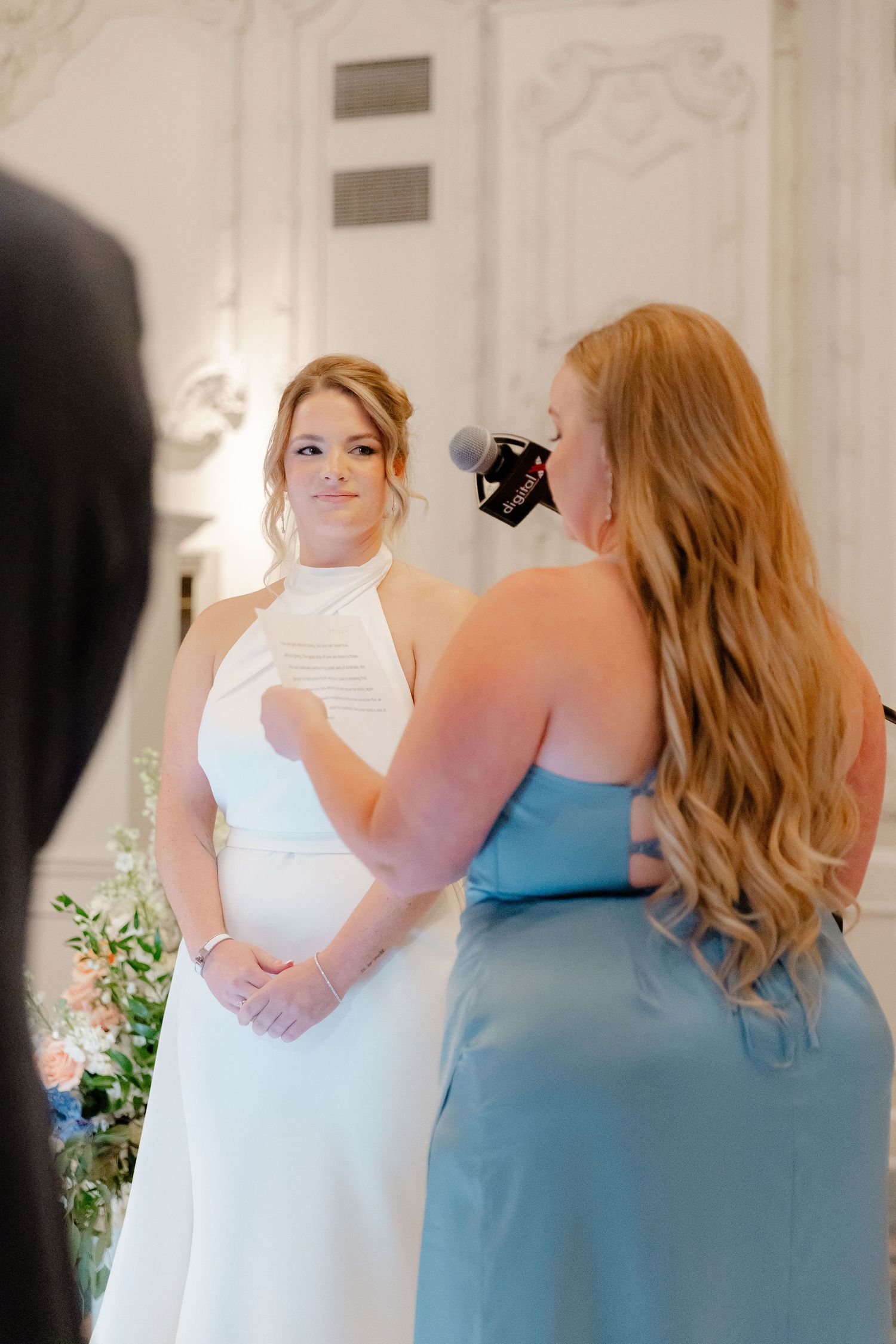 Two women in white and blue dresses share an emotional moment during a wedding celebration.