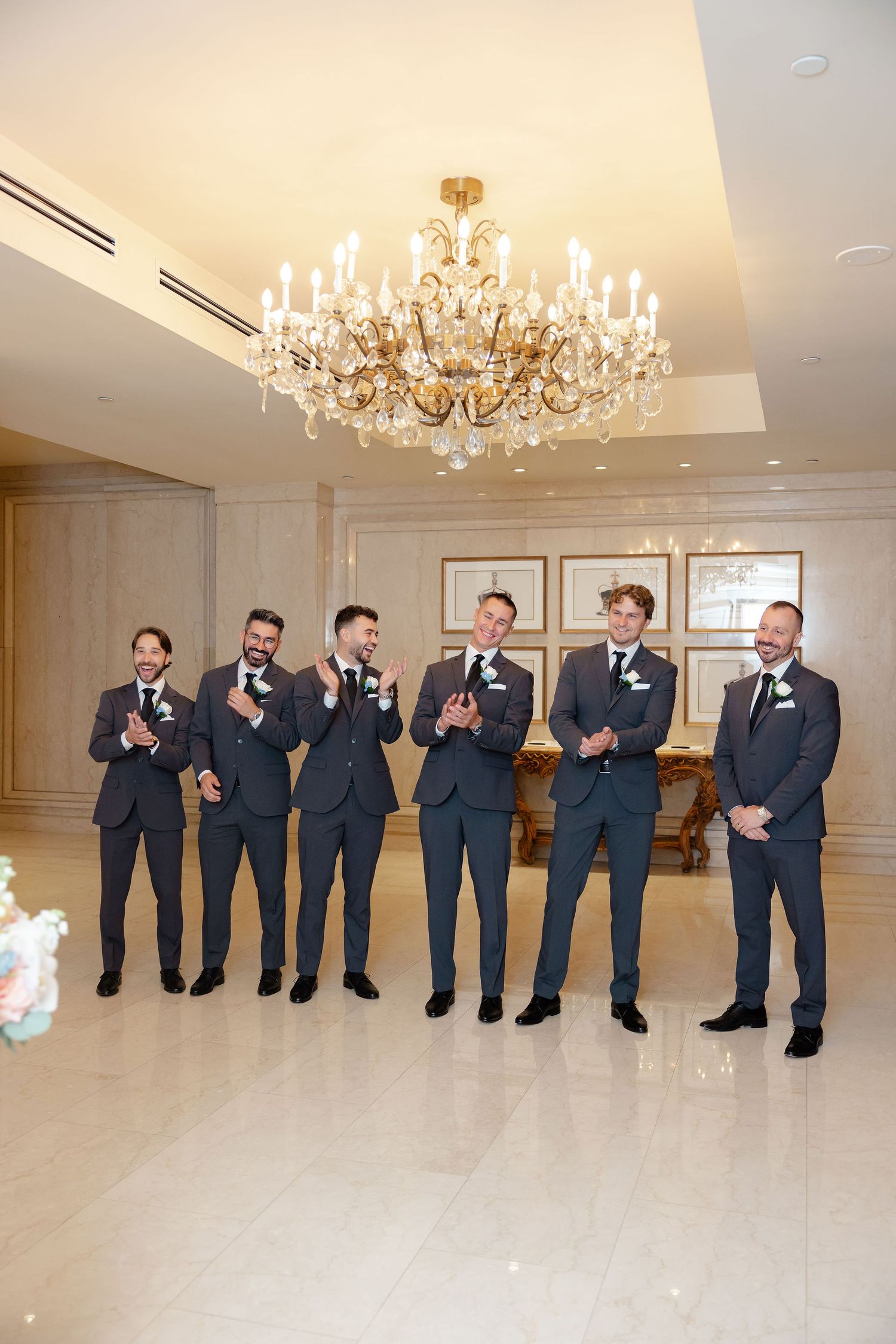 Groomsmen in dark suits pose playfully in a line under crystal chandeliers in an elegant ballroom.