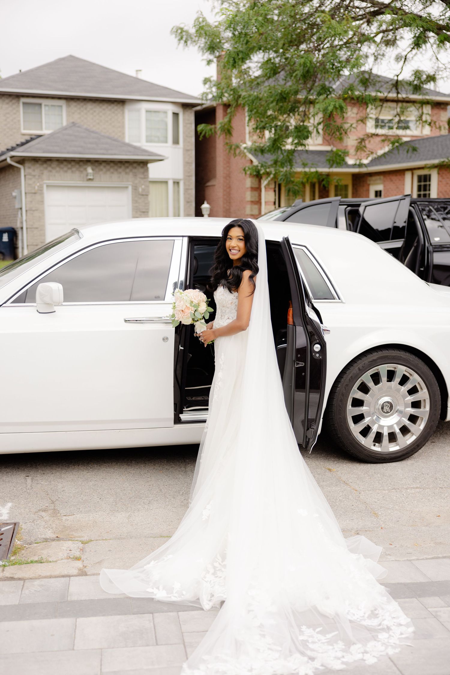 Bride in white lace gown steps out of luxury white car holding bouquet.