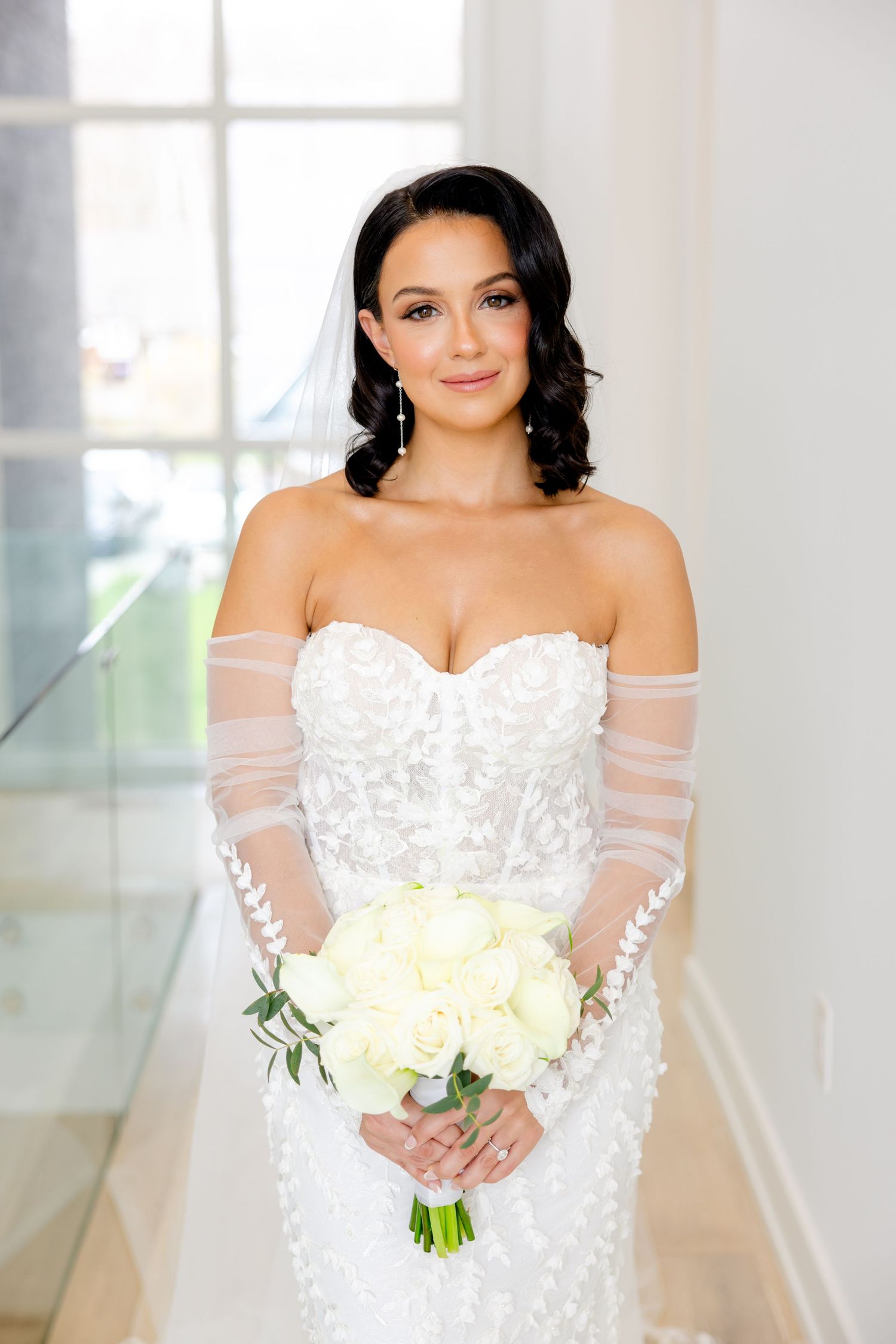 Close-up portrait of bride in off-shoulder lace wedding dress holding cream roses in modern white hallway.